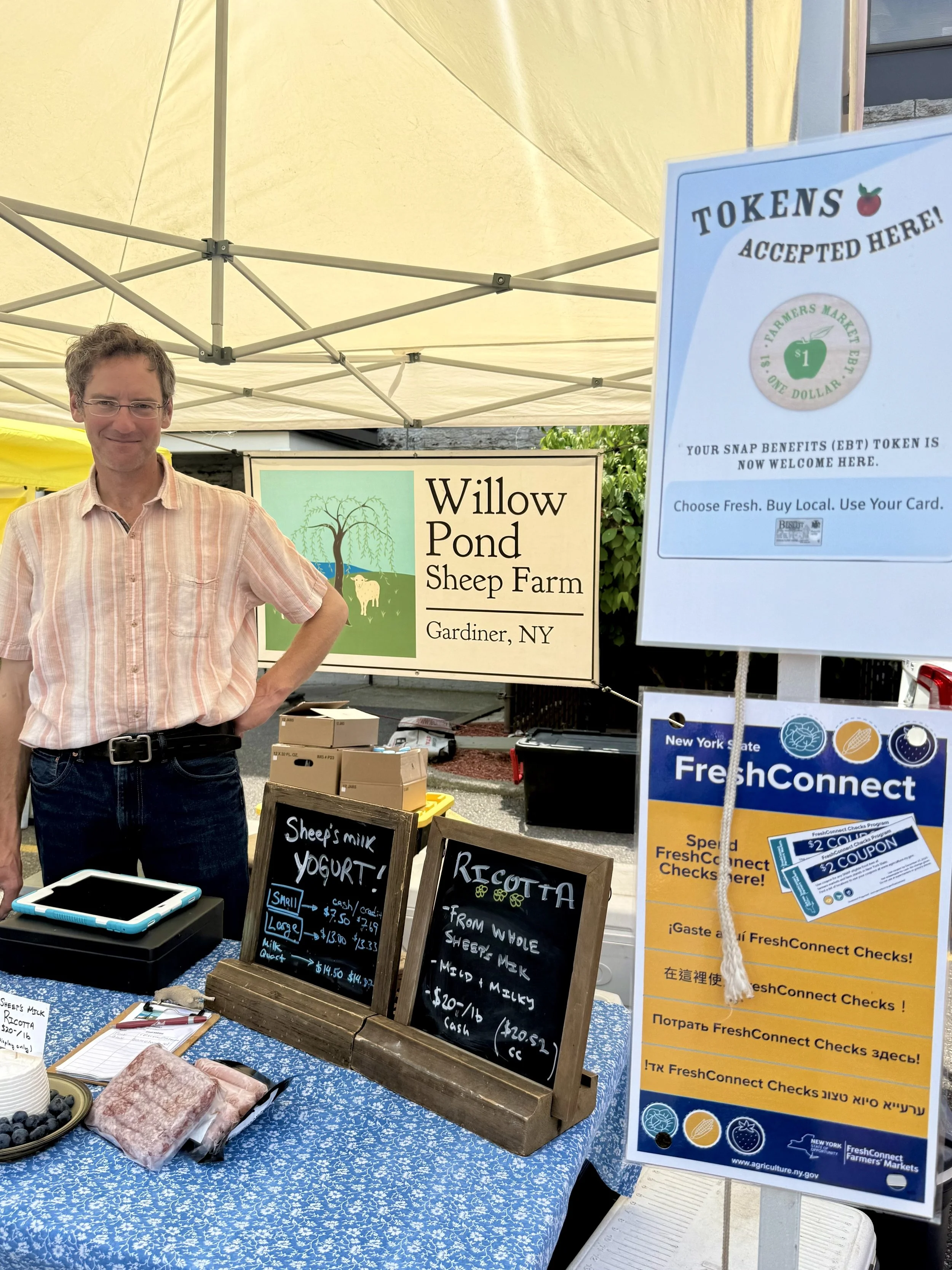 A man standing behind a table at a farmers' market stall. The table has a blue patterned cloth, and several chalkboards with handwritten menus for sheep's milk yogurt and ricotta cheese are displayed. There are small containers of blueberries and blo