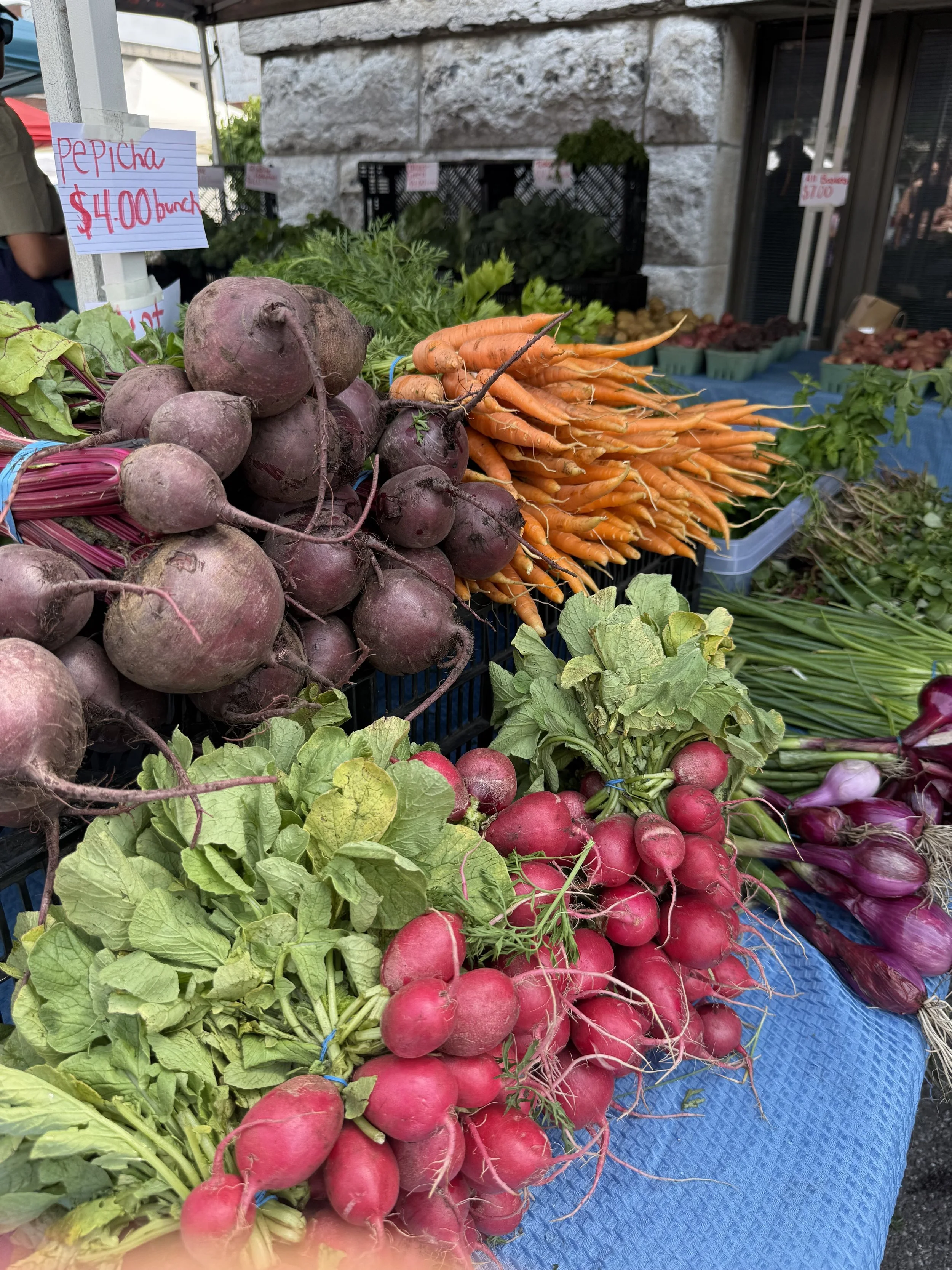 Fresh vegetables displayed at a market stand, including a bunch of purple beets, orange carrots, and red radishes with green leaves, on a blue tablecloth.