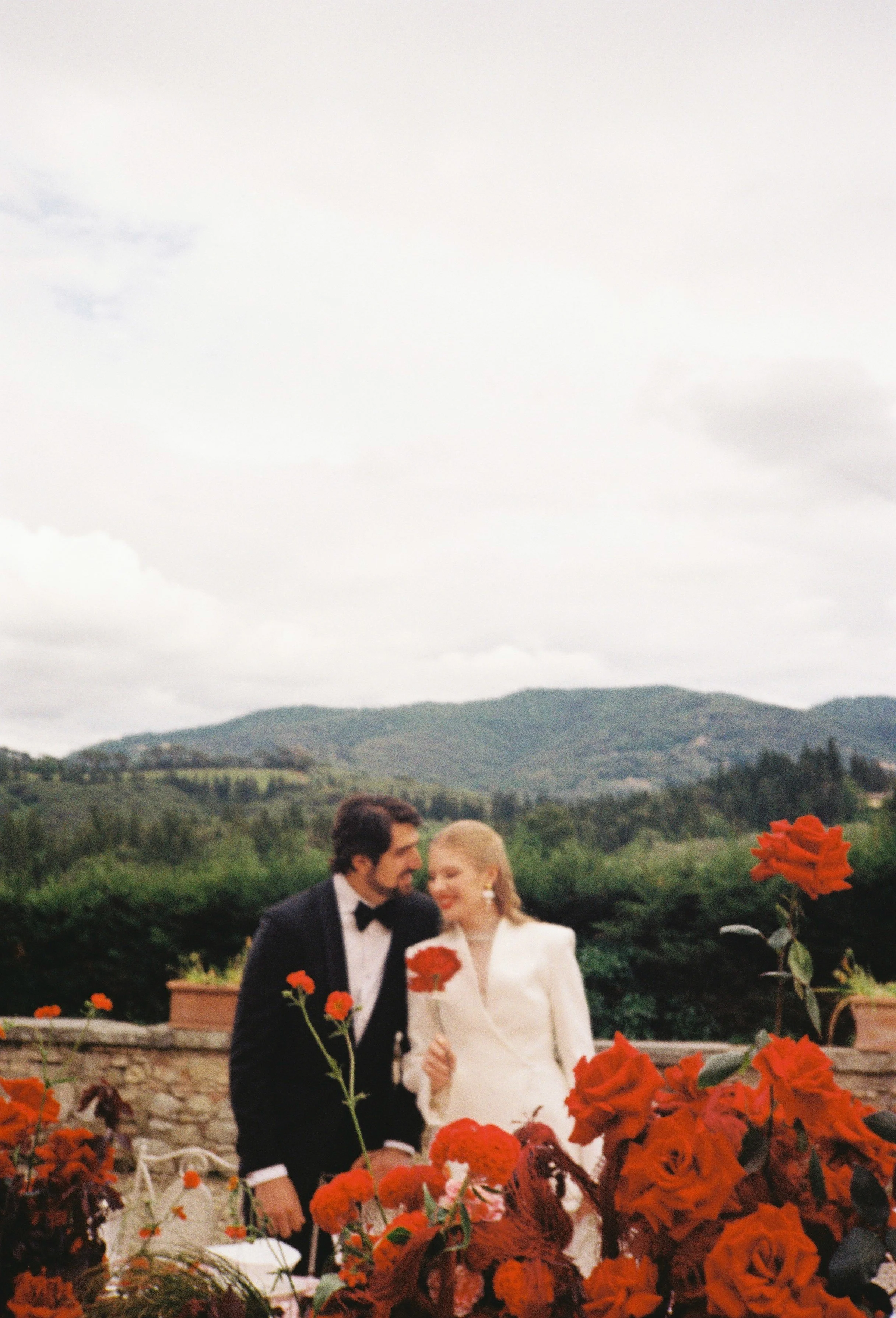 Film photography editorial wedding portrait in the Tuscan countryside, Italy by Cleya Asulon, destination wedding photographer for Tuscany and Italian destinations — bride and groom seated at an outdoor reception table on a stone terrace overlooking 
