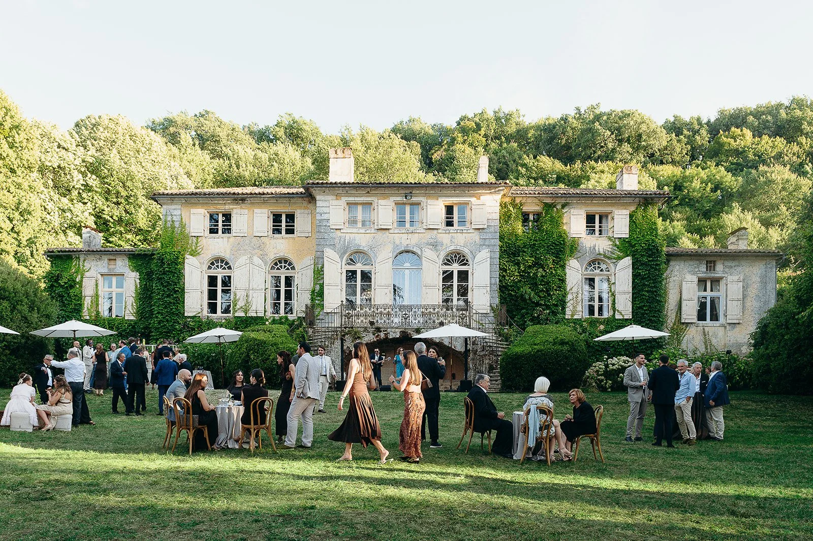 Outdoor wedding reception in front of historic mansion Domaine de Perrotin, in Gironde, south west of France, with guests seated and socializing under umbrellas on a grassy lawn surrounded by trees.