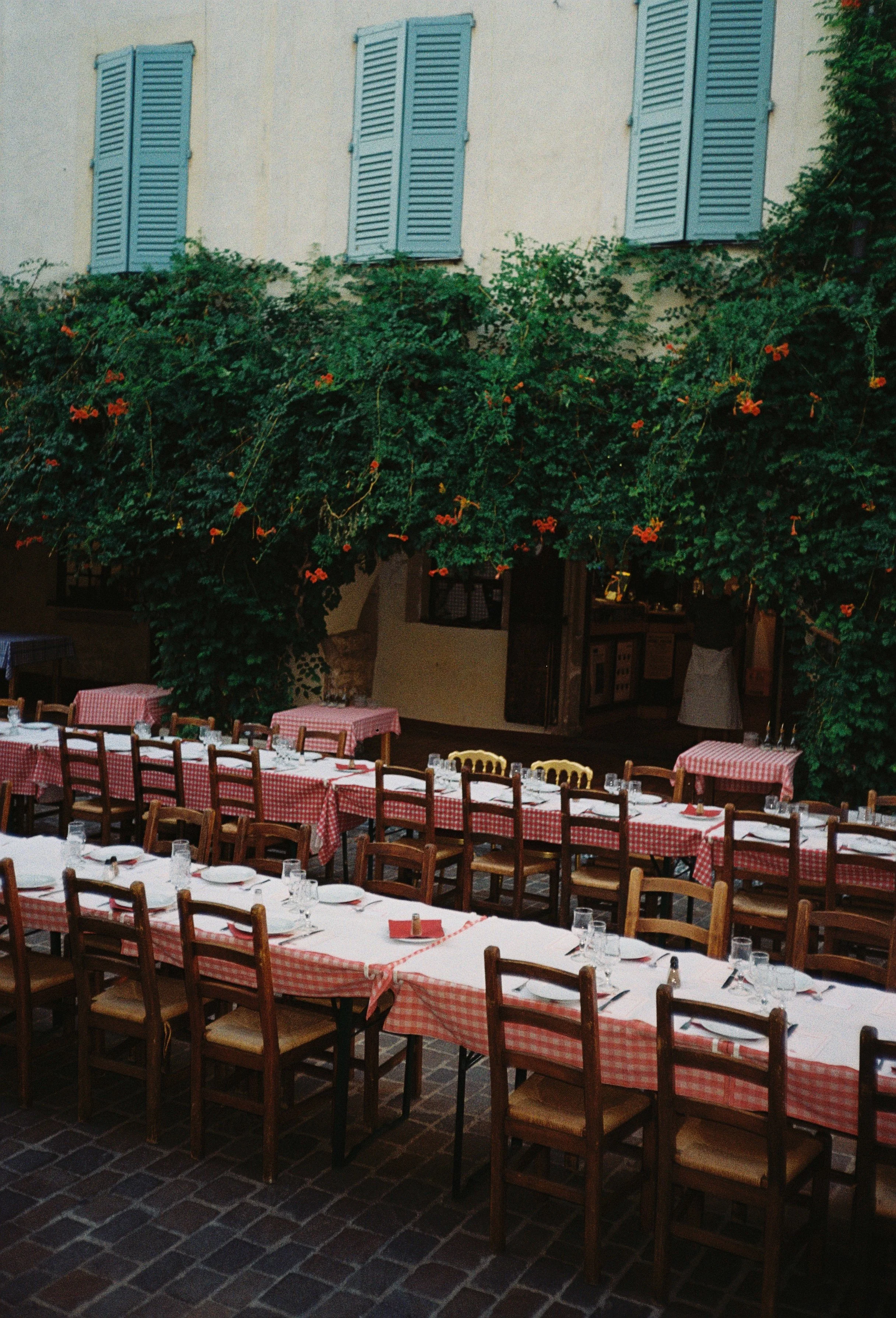 Film photography editorial wedding reception setup in the heart of Biot village on the French Riviera by Cleya Asulon, destination wedding photographer for the Côte d'Azur — long wooden banquet tables dressed with classic red gingham tablecloths, sim