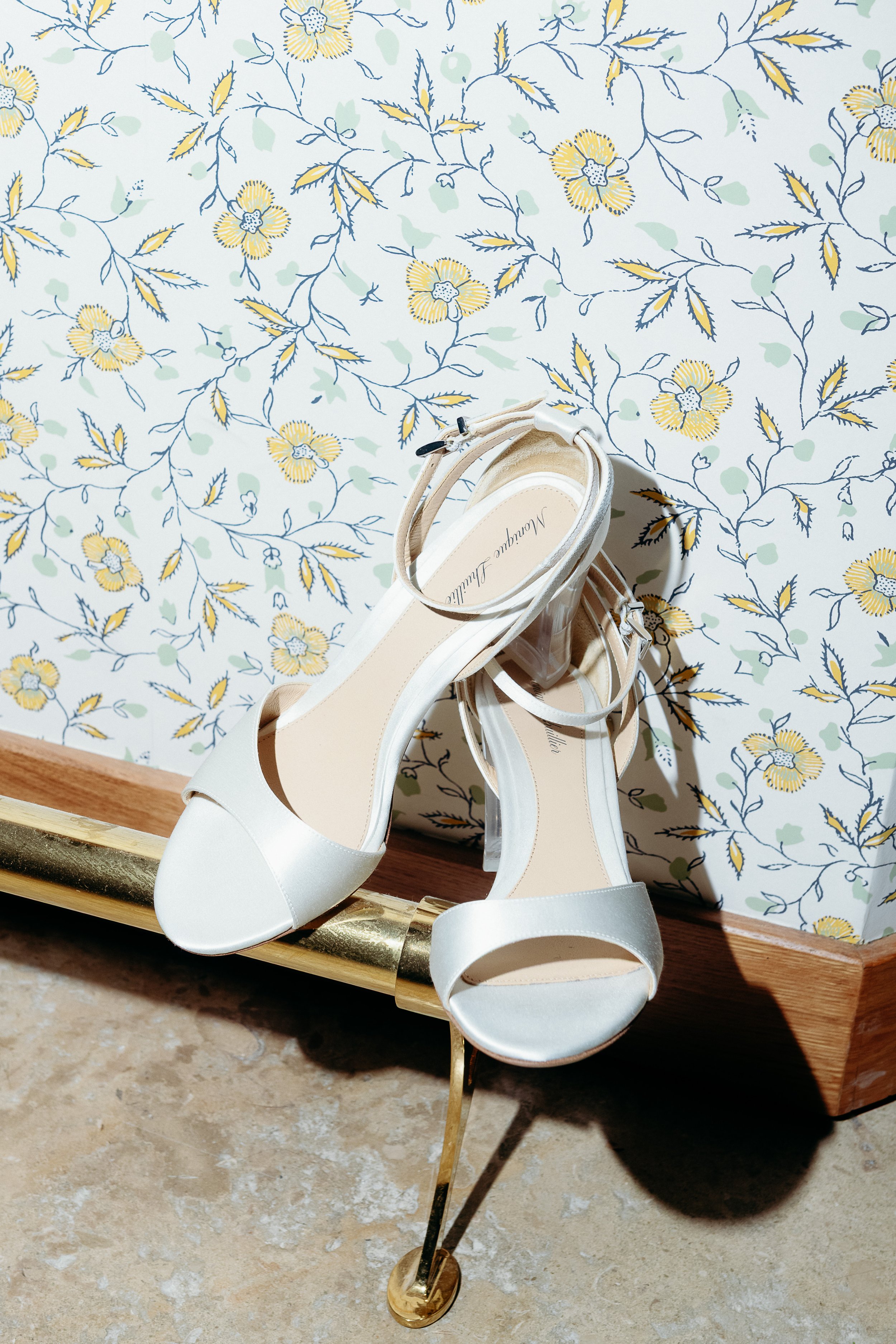 Bridal detail shot at Domaine de Fontenille in Provence: a pair of ivory satin Monique Lhuillier wedding sandals with ankle straps and transparent block heels, resting on a gold-trimmed wooden console against a yellow and blue floral block-print wall