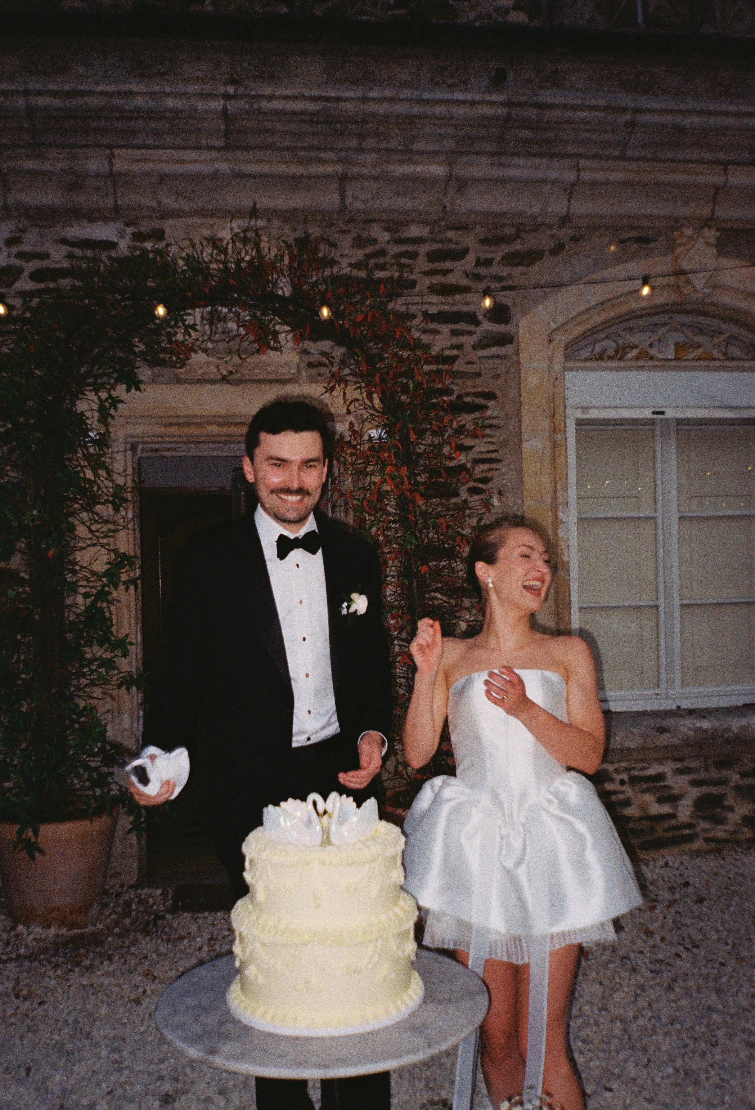 Film photography editorial wedding cake-cutting moment at a private château in the Dordogne by Cleya Asulon, destination wedding photographer for the Dordogne and Périgord region — bride and groom captured in a joyful candid moment beside a two-tier 