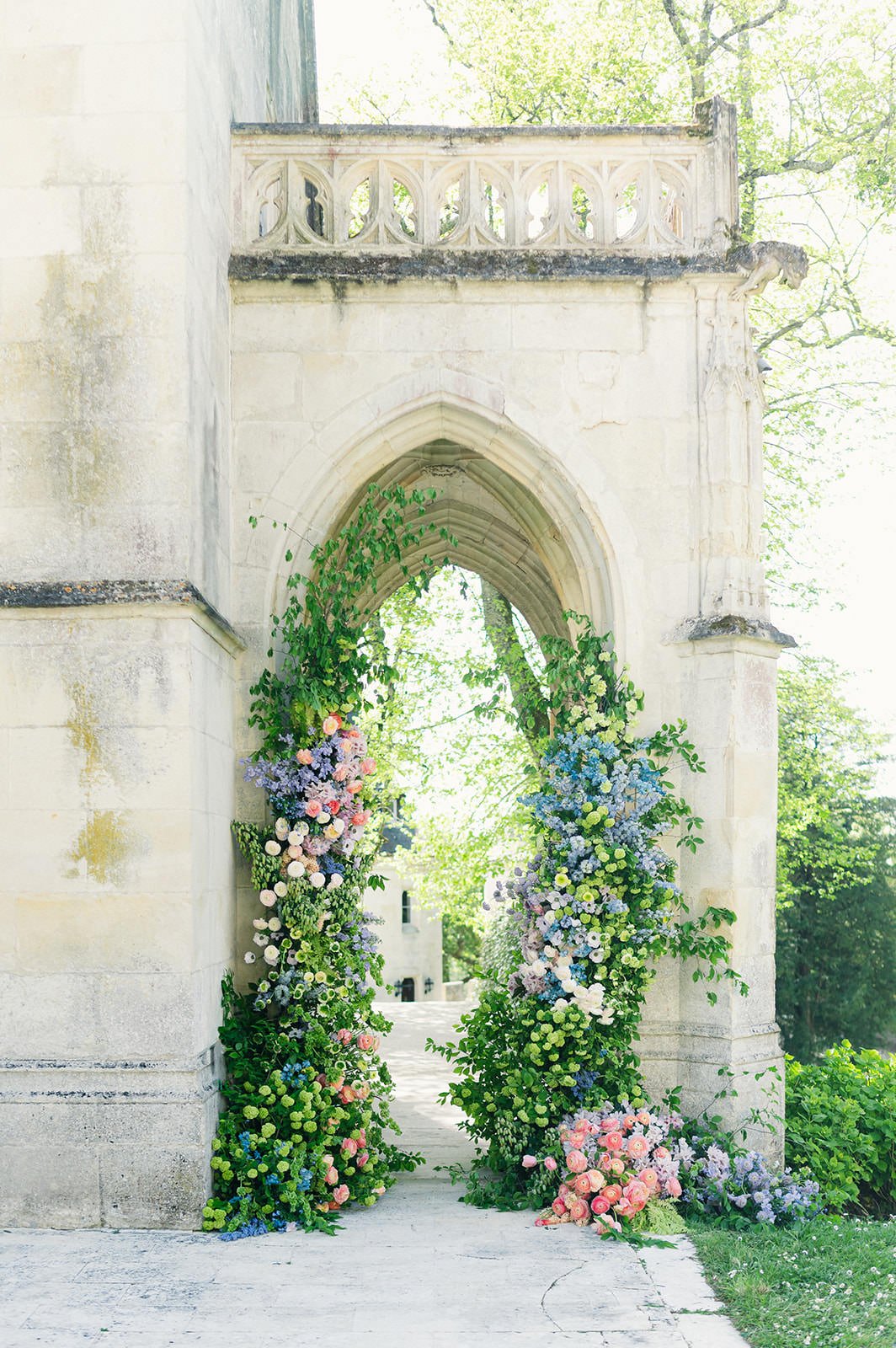 Romantic ceremony floral installation at Château de Mirambeau (Gironde, France) by Cleya Asulon, destination wedding photographer based in Bordeaux and Cognac — neo-Gothic stone archway with carved tracery and gargoyle framing a lush asymmetrical flo
