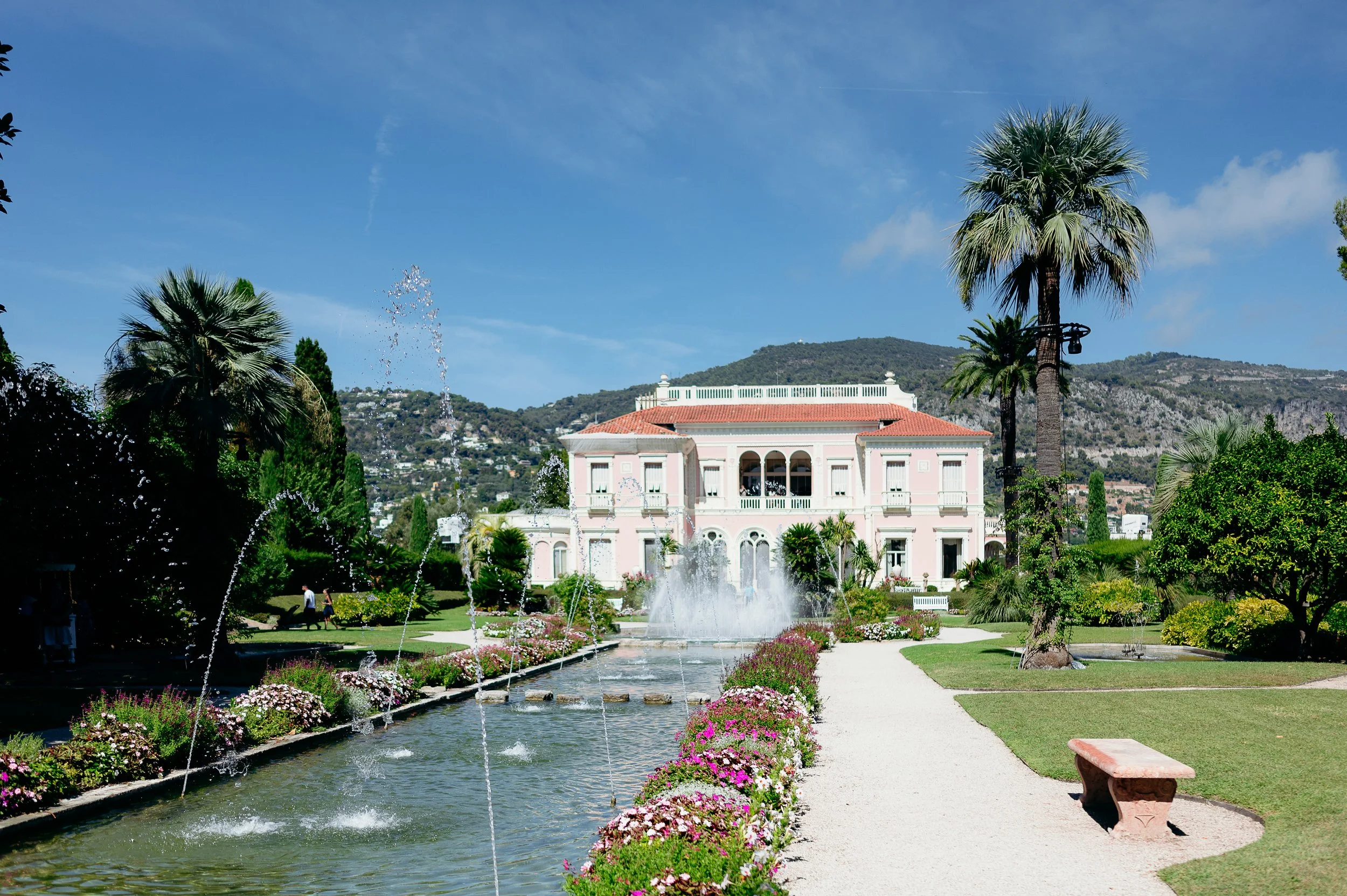View of the pink Belle Époque façade of Villa Ephrussi de Rothschild in Saint-Jean-Cap-Ferrat, French Riviera, seen from the main garden axis with its long reflecting pool, dancing water jets, pink and white flowerbeds, palm trees, gravel pathway and