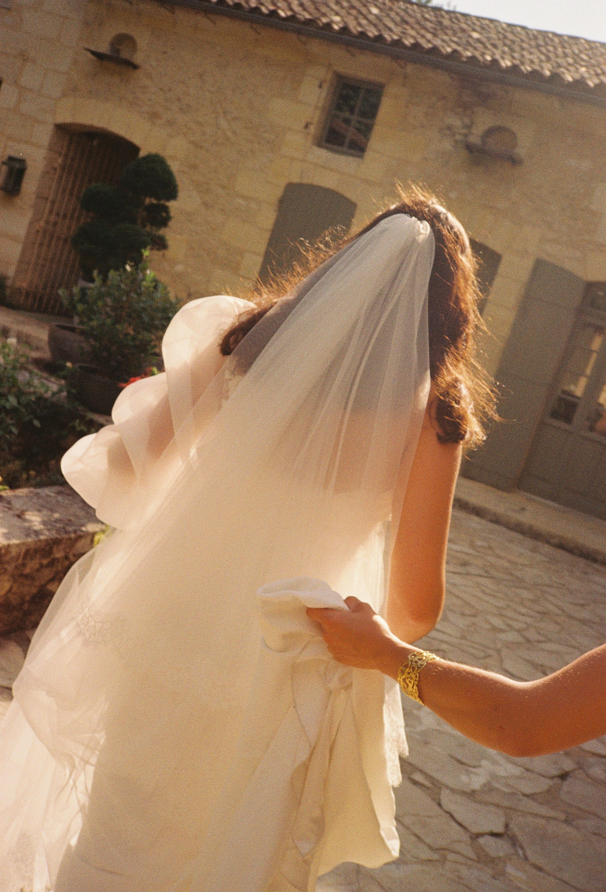 Film photography editorial bridal portrait at Domaine de Perrotin near Bordeaux, France by Cleya Asulon, destination wedding photographer for Bordeaux and the Gironde region — bride captured in motion walking across a sunlit cobblestone courtyard, we