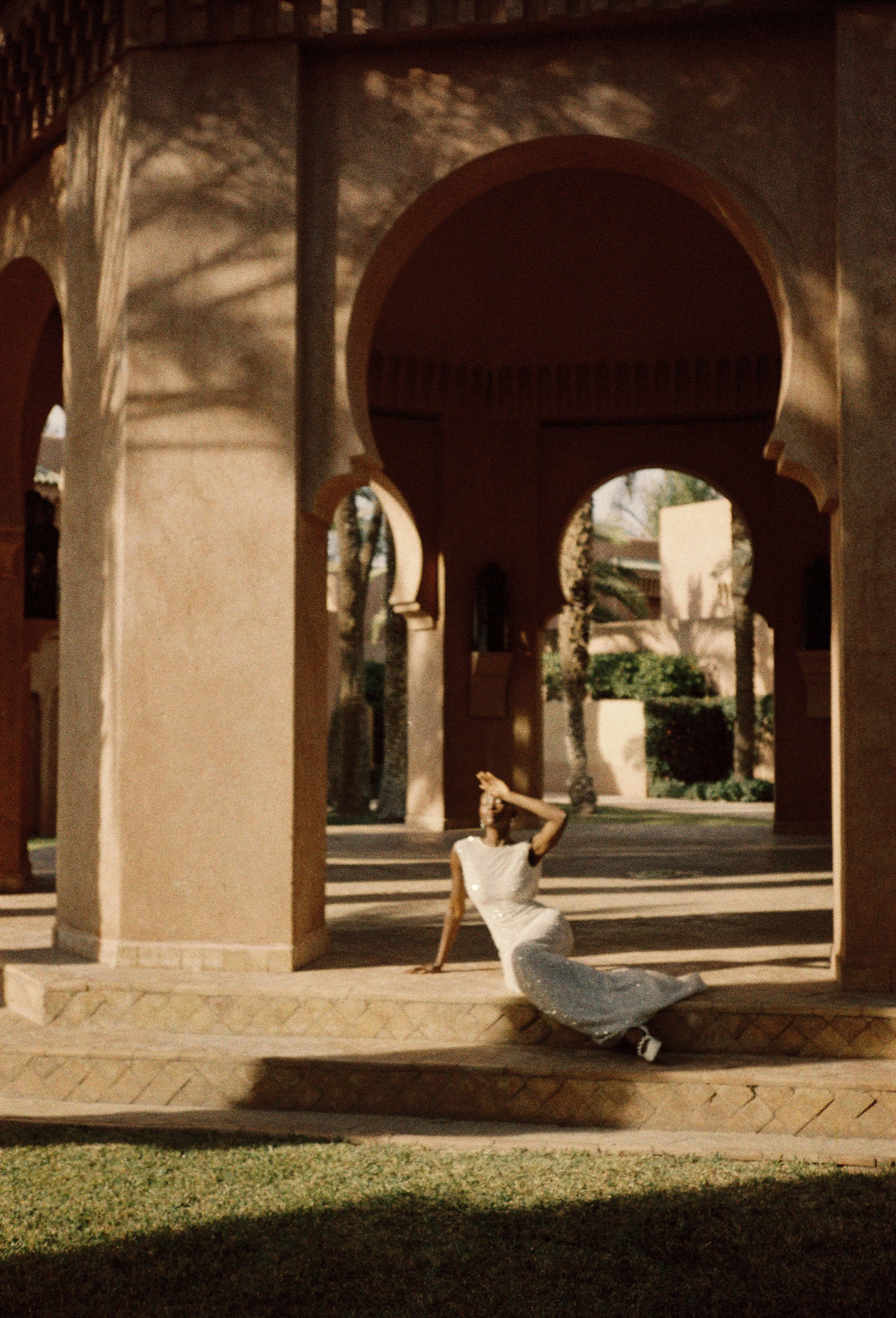Film photography editorial bridal portrait at Amanjena resort in Marrakech, Morocco by Cleya Asulon, destination wedding photographer for Marrakech and Moroccan destinations — bride reclining gracefully on a tiled step beneath monumental Moorish hors