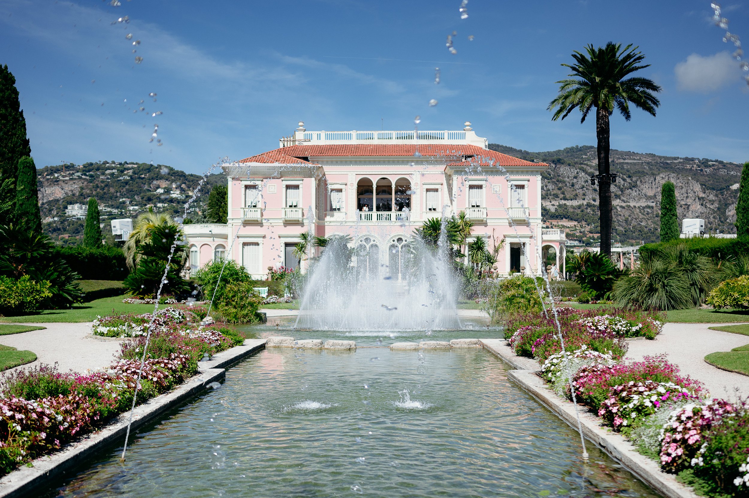 Symmetrical view of the pink Belle Époque façade of Villa Ephrussi de Rothschild in Saint-Jean-Cap-Ferrat, French Riviera, framed by the central reflecting pool and its grand fountain in full spray, flanked by parterres of pink and white flowers, pal