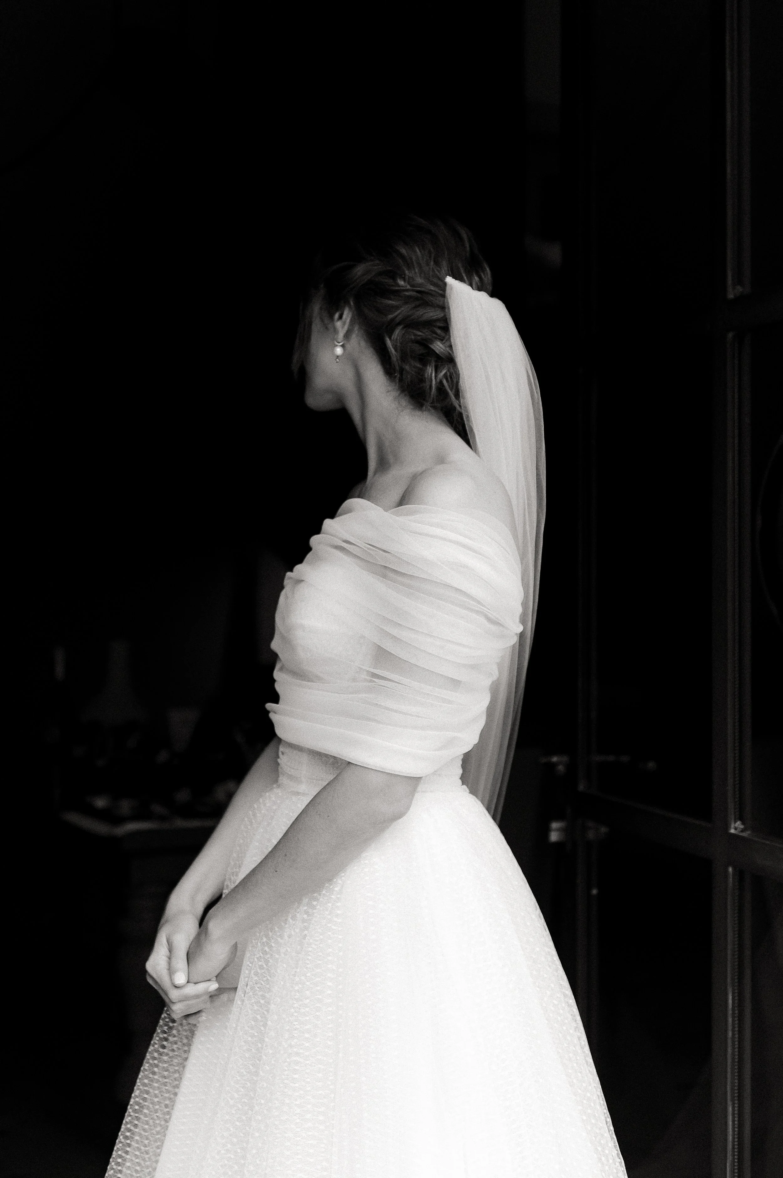 Black and white editorial portrait of a bride at Domaine de Fontenille in Provence, photographed in profile against a dark interior with natural window light, wearing an off-the-shoulder draped tulle Monique Lhuillier wedding gown with a soft updo, p
