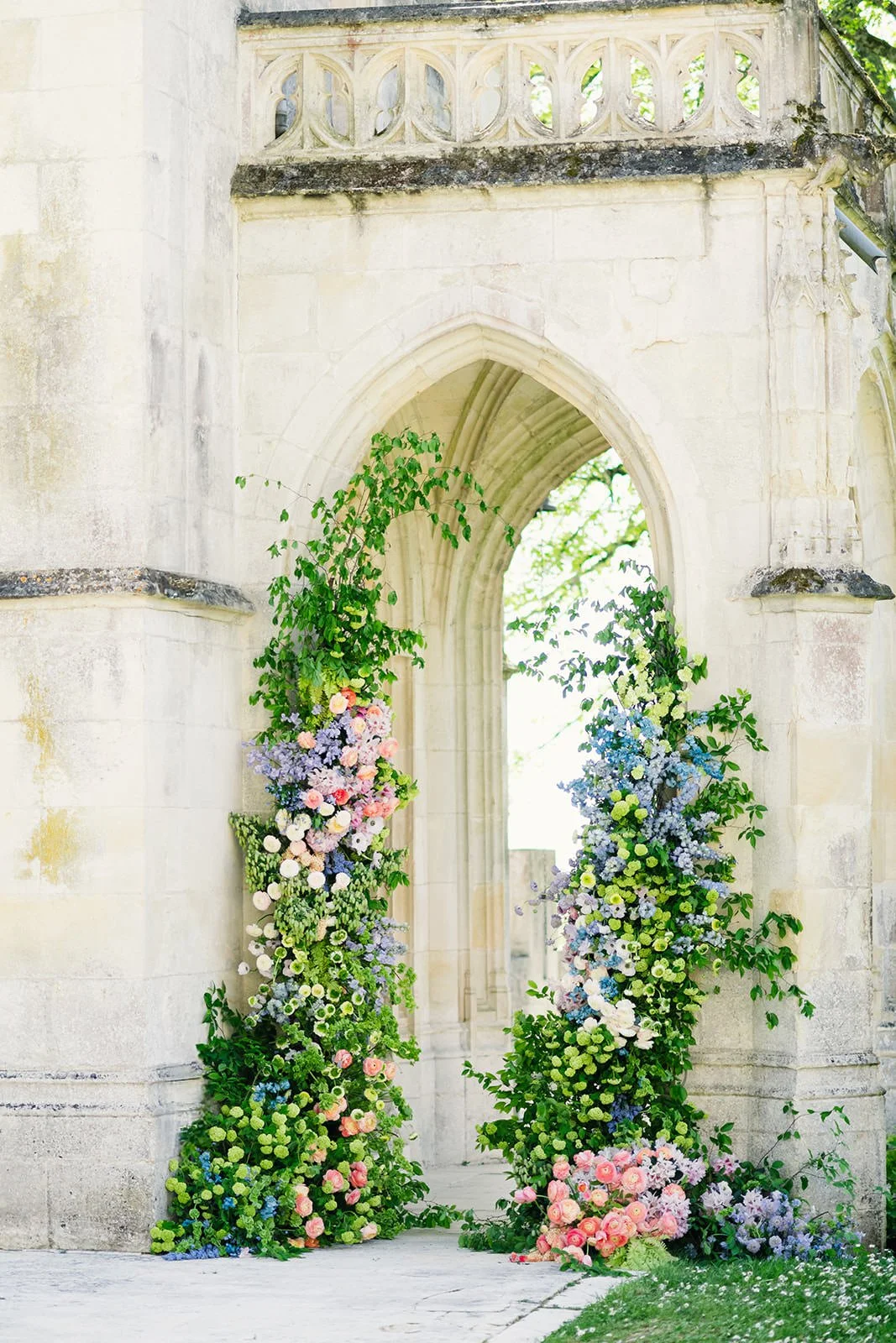 Romantic ceremony floral installation at Château de Mirambeau (Gironde, France) by Cleya Asulon, destination wedding photographer based in Bordeaux and Cognac — neo-Gothic stone archway with carved tracery framing a lush asymmetrical pastel floral ar