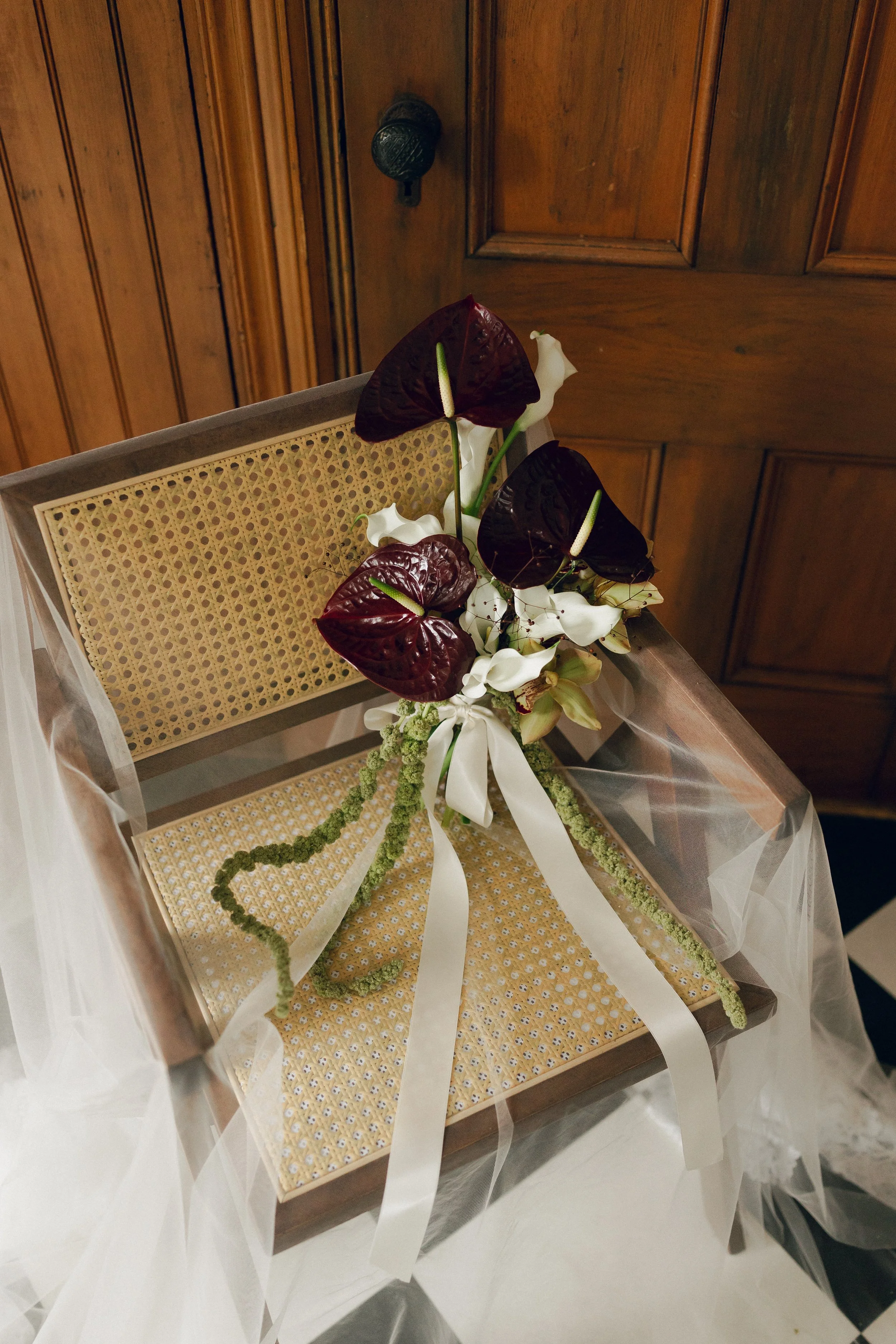A floral arrangement with dark purple and white calla lilies and anthuriums, tied with a white ribbon, resting on a woven cane chair draped with sheer fabric, against wooden paneled walls.