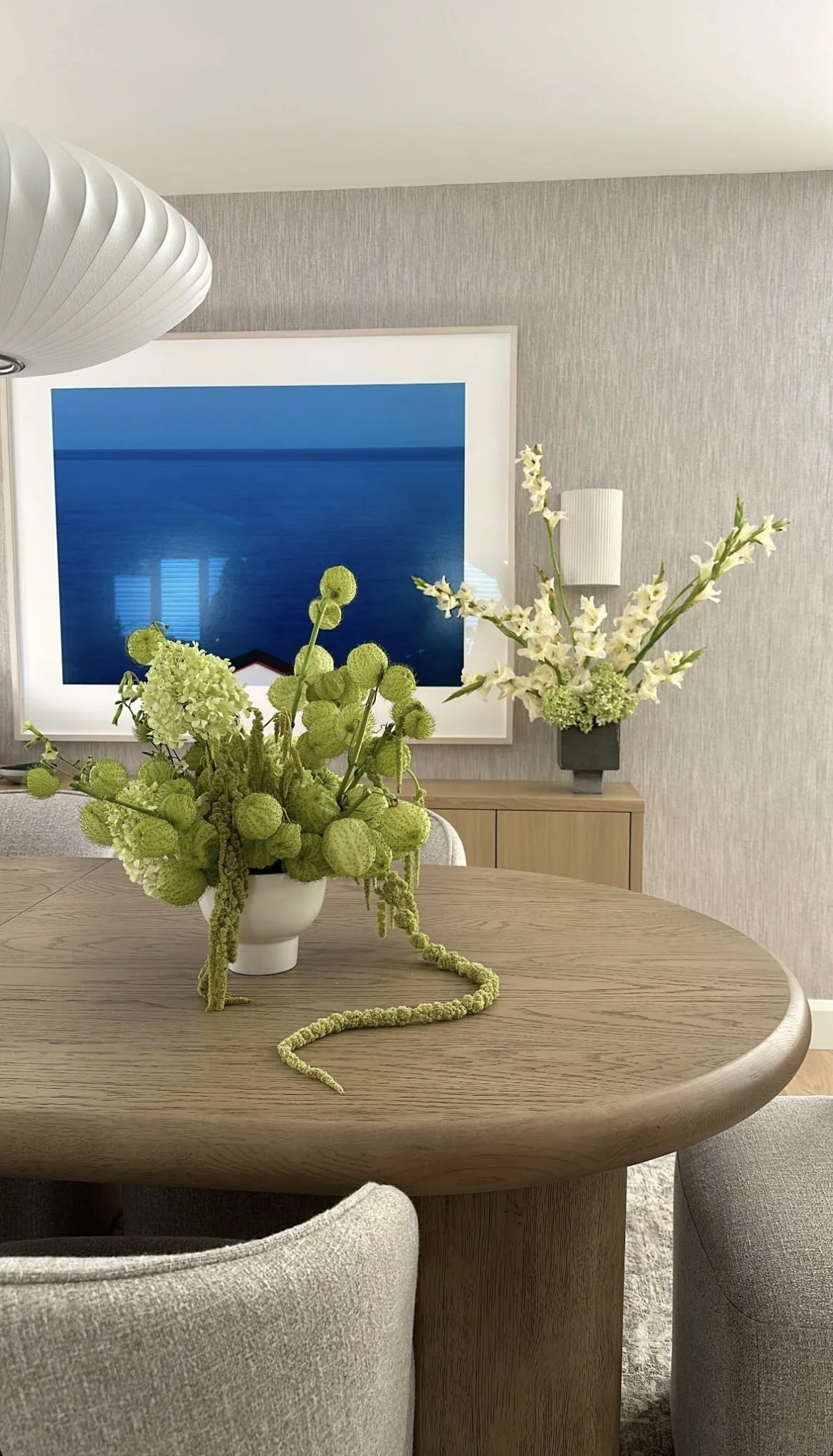 A contemporary dining room features a wooden table with two light gray upholstered chairs. A white vase with green, textured foliage and flowers sits on the table. In the background, there is a framed blue ocean photograph, a wooden sideboard, and a modern wall sconce on a textured beige wall.