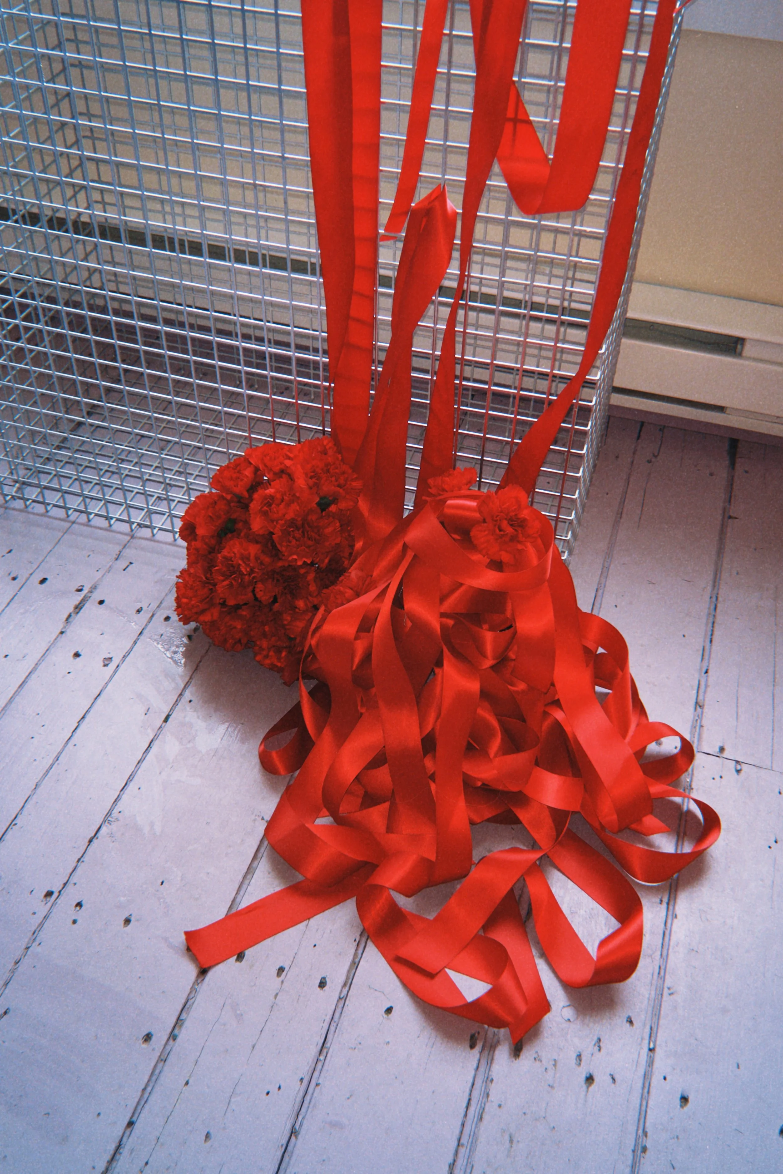 A bouquet of red carnations with long red ribbons, placed on a wooden floor next to a metal wire fence.
