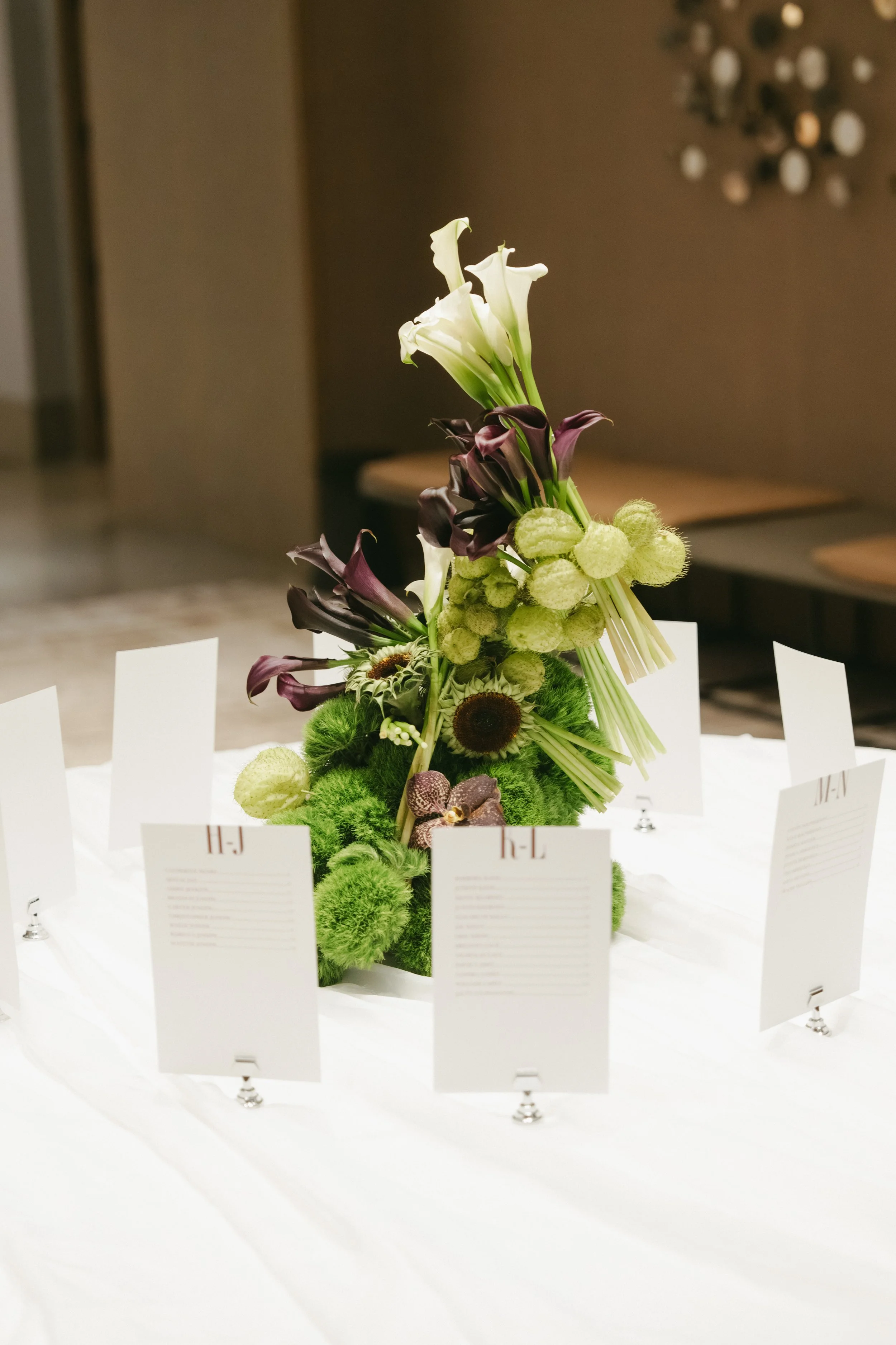 Elegant flower centerpiece with white calla lilies, purple calla lilies, green moss, and other flowers, surrounded by small white cards on a white tablecloth.
