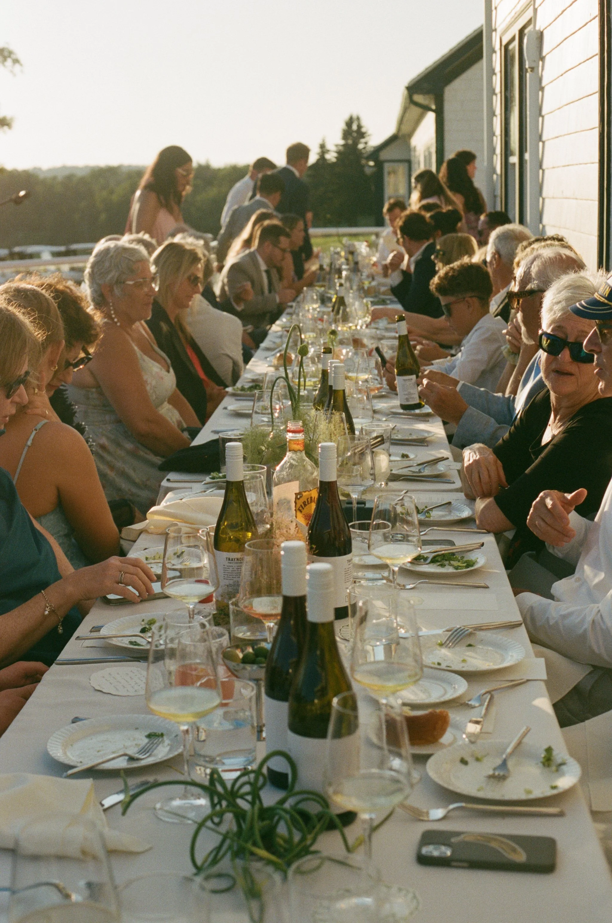 People gathered around a long outdoor dining table with wine bottles, glasses, and plates during sunset.