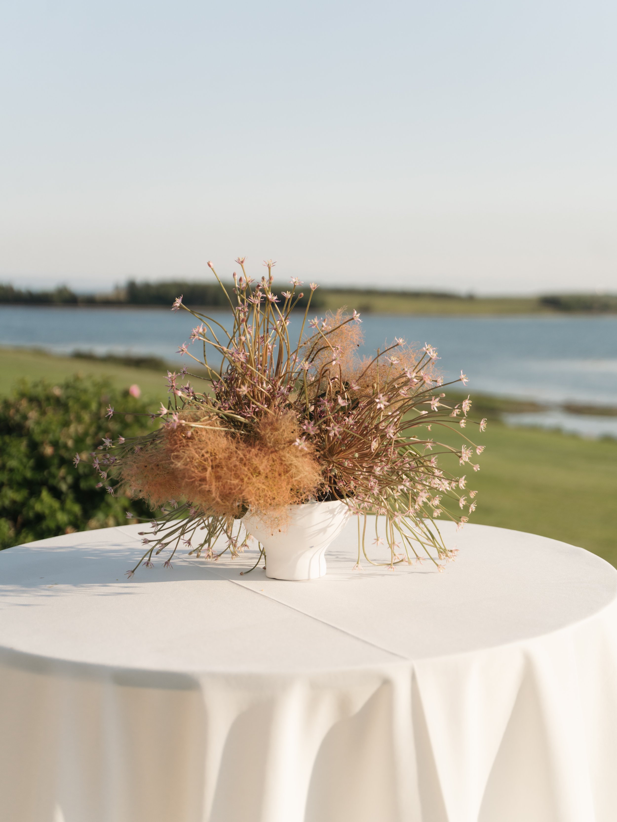 A white vase with pink and brown dried flowers on a white round table beside a body of water with a grassy shoreline and distant trees, under a clear blue sky.