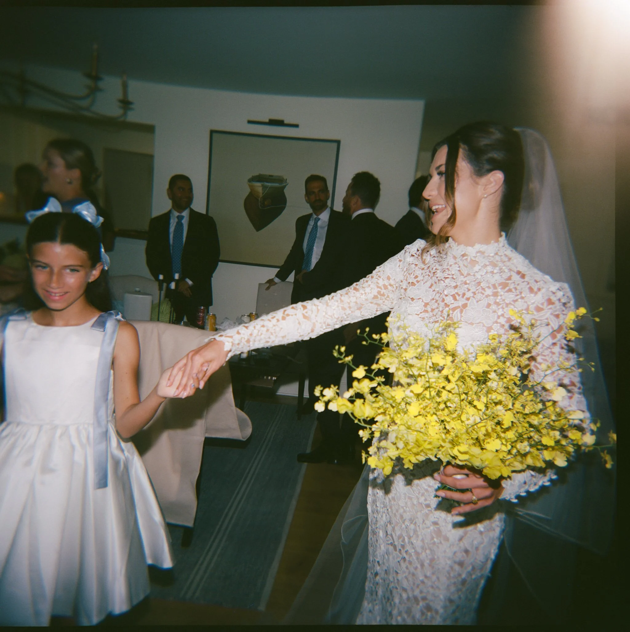 A bride in a white lace wedding dress holding a yellow bouquet walks hand-in-hand with a young girl in a white dress, while guests in formal wear look on.
