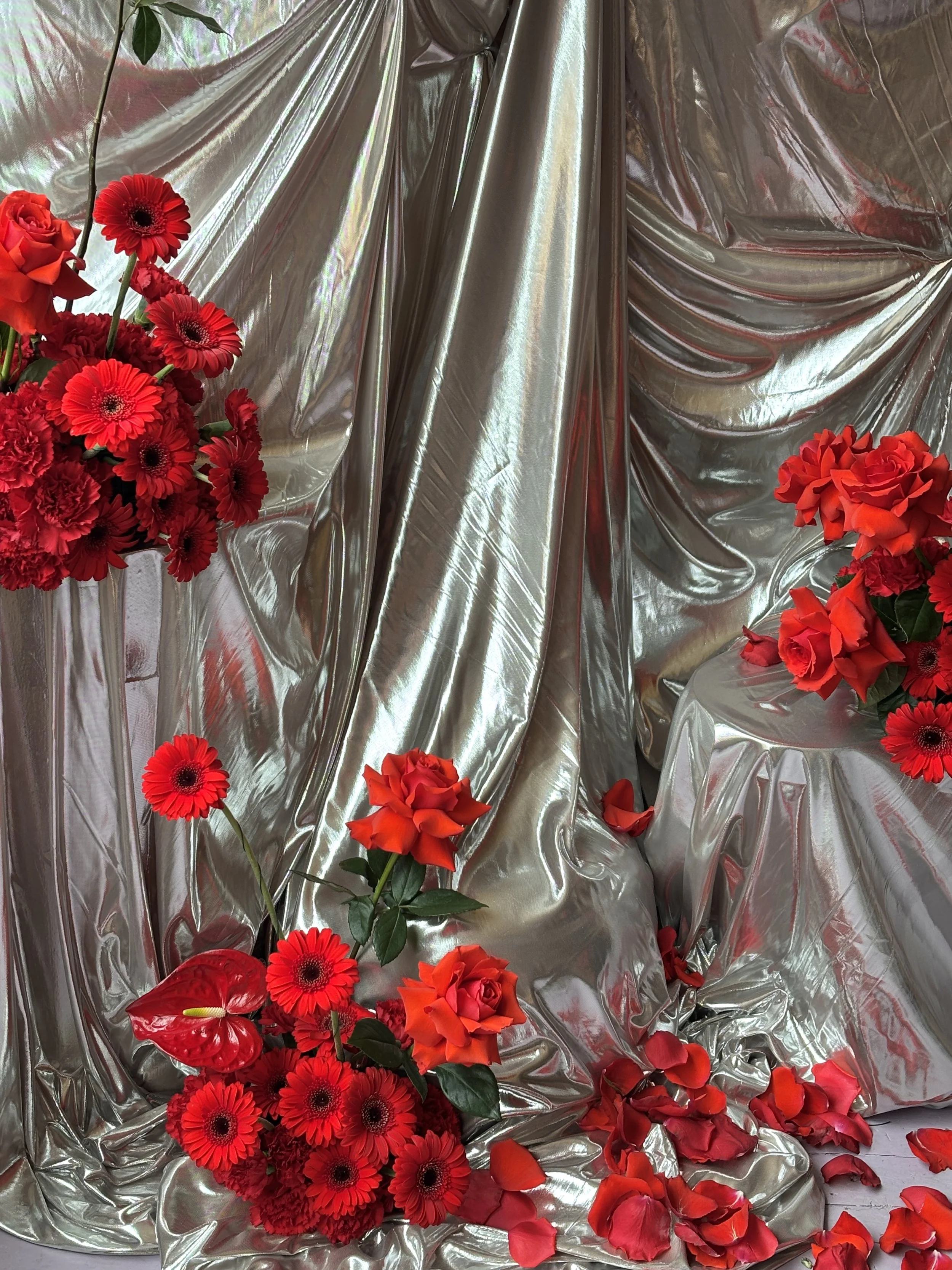 Red flowers, including roses, gerbera daisies, and anthuriums, arranged around shiny silver fabric backdrop with some rose petals scattered on the ground.
