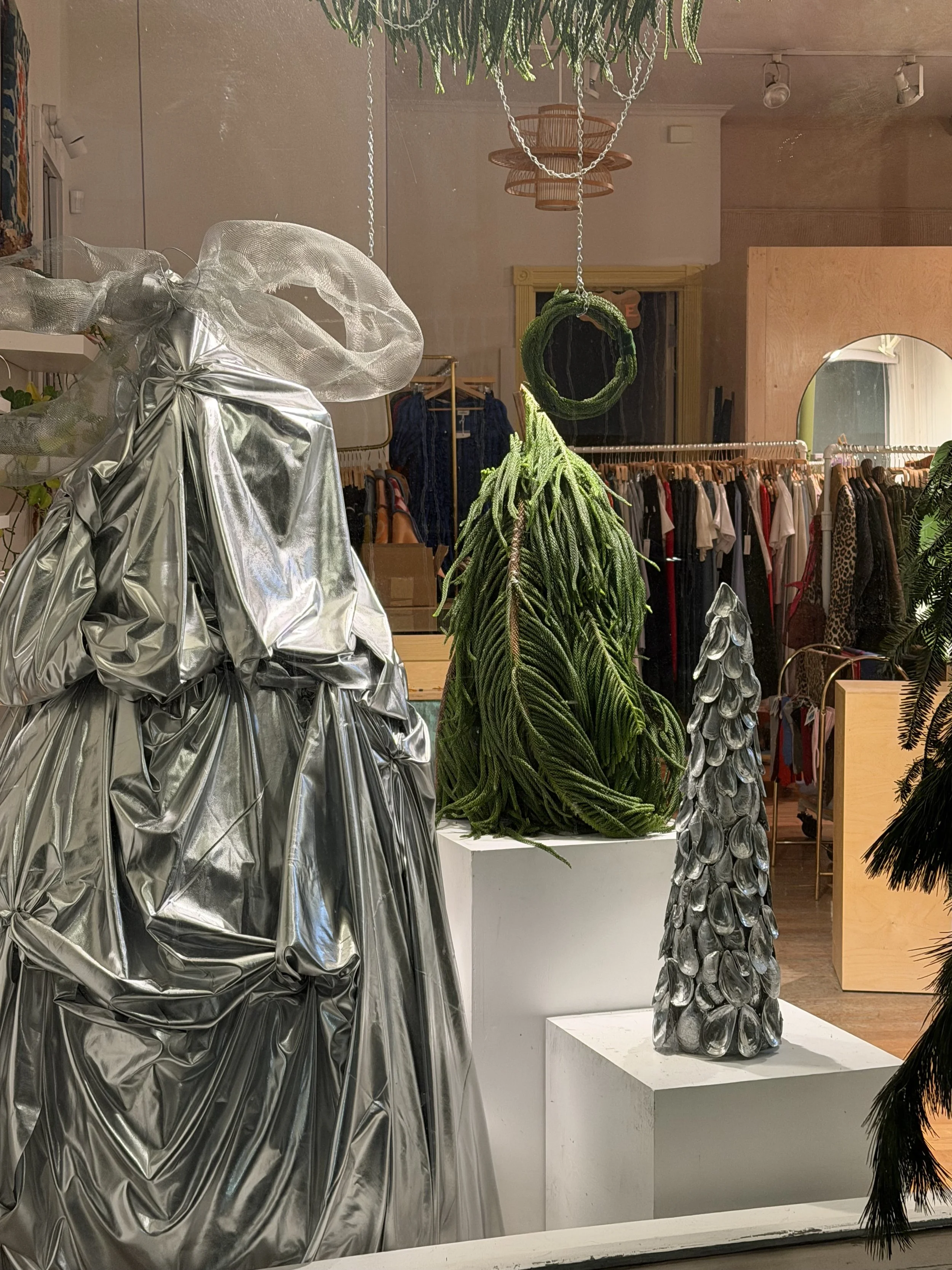 Store window display with a metallic silver, ruffled dress, a green hanging plant, and a silver cone-shaped sculpture resembling a Christmas tree.