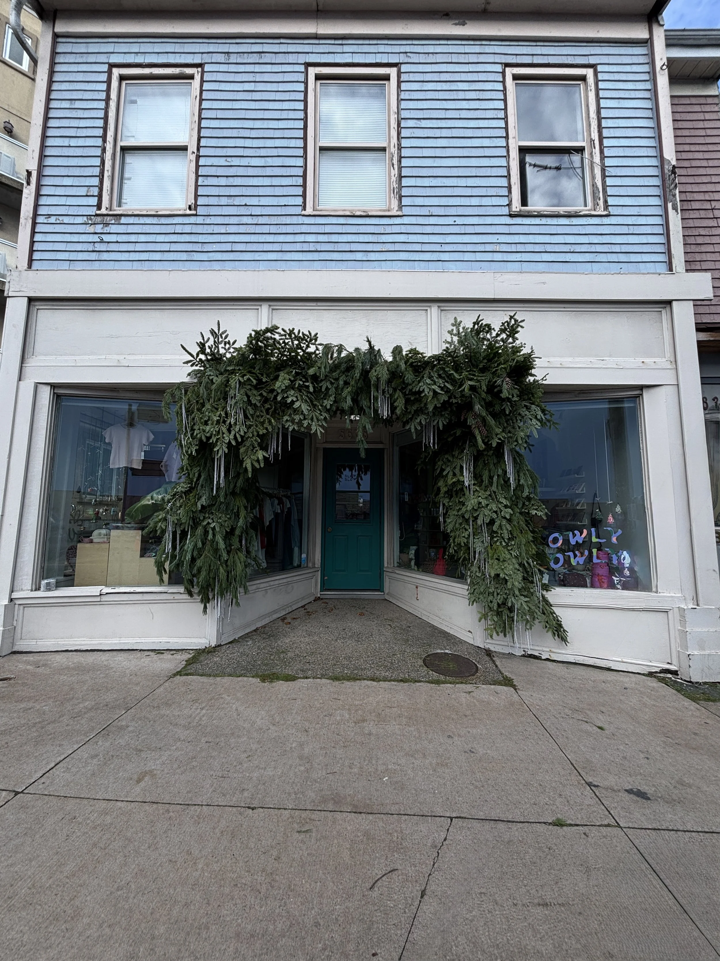 Storefront decorated with green pine branches and icicles above the entrance, featuring large display windows and a teal door, with a blue, weathered two-story building facade.