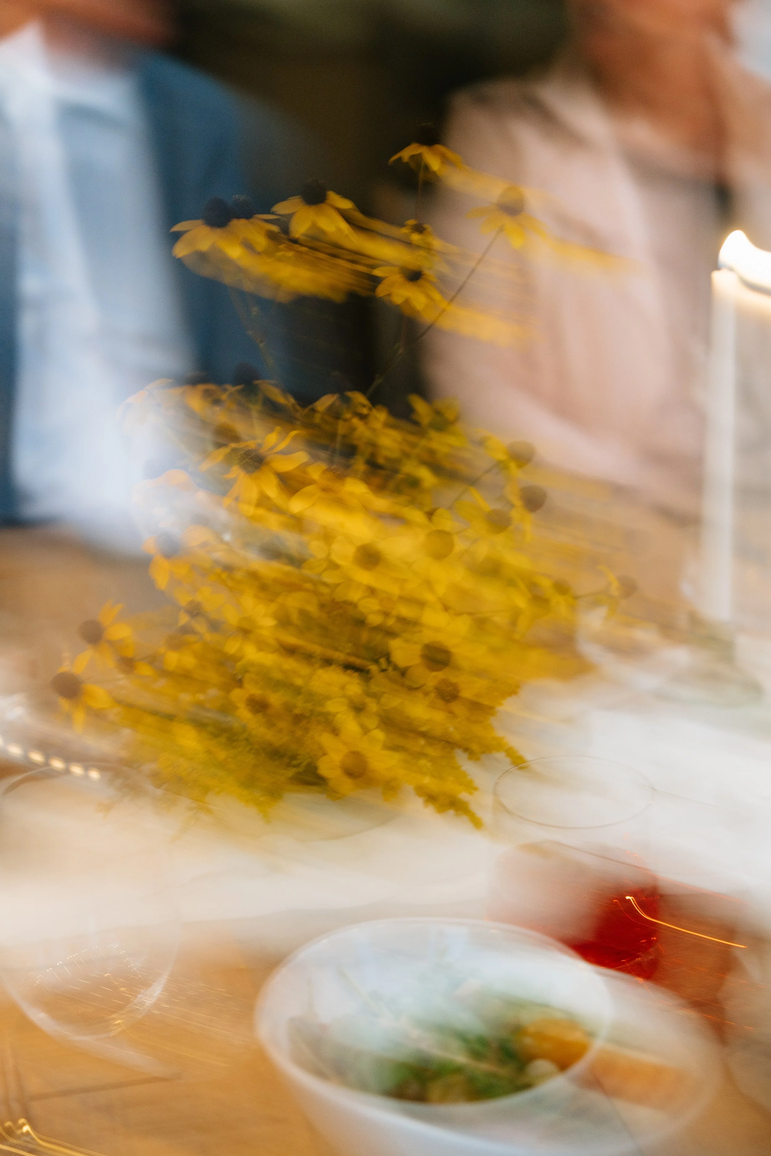 Blurred image of a table with a bouquet of yellow flowers, a white bowl with food, and two people sitting nearby, with motion blur and abstract lighting.