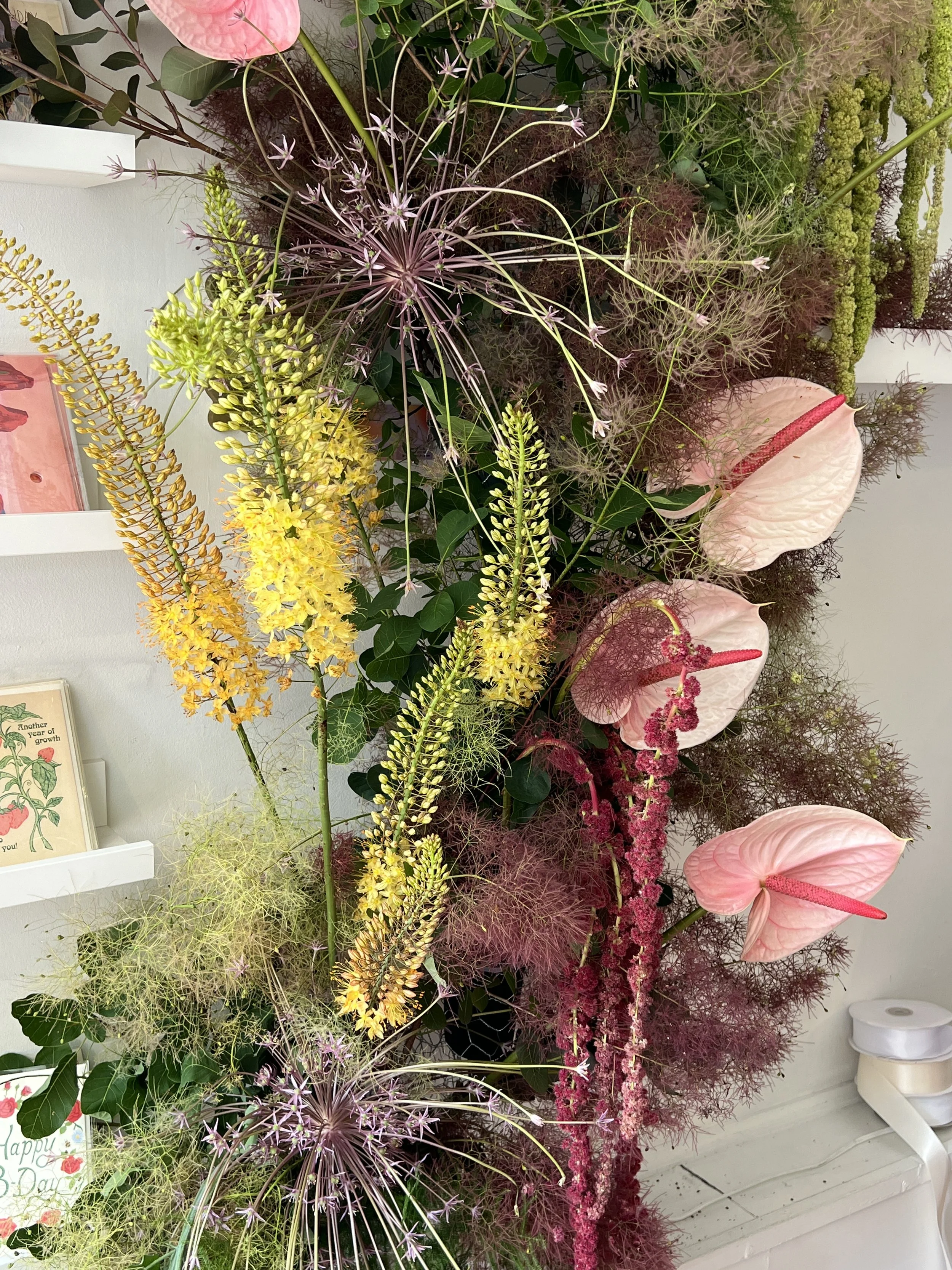 A colorful floral display with pink anthuriums, yellow and purple flowers, and green foliage arranged indoors.