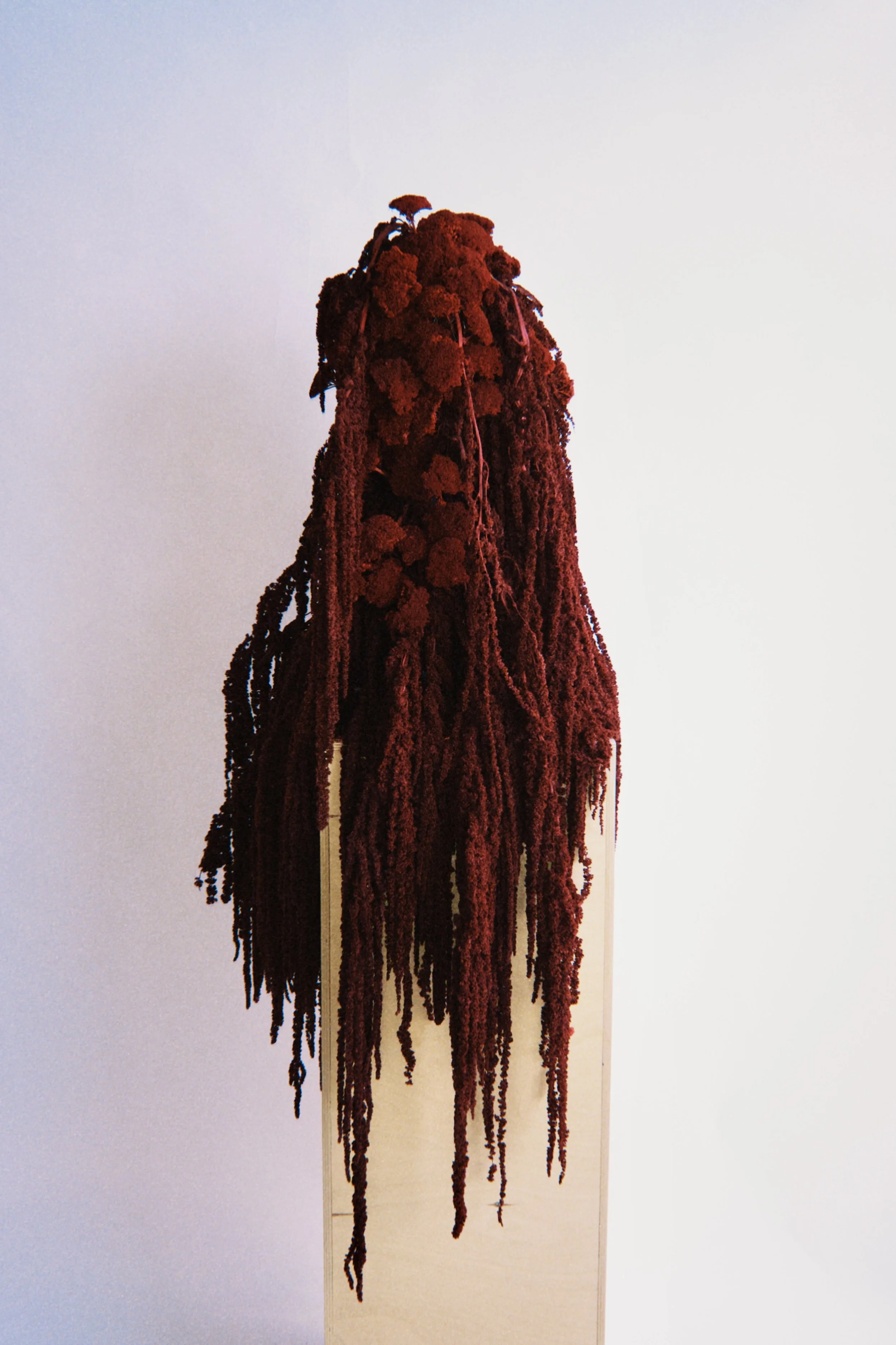 A textured art piece resembling a head with long, red, yarn-like strands flowing downward, displayed on a stand against a plain background.
