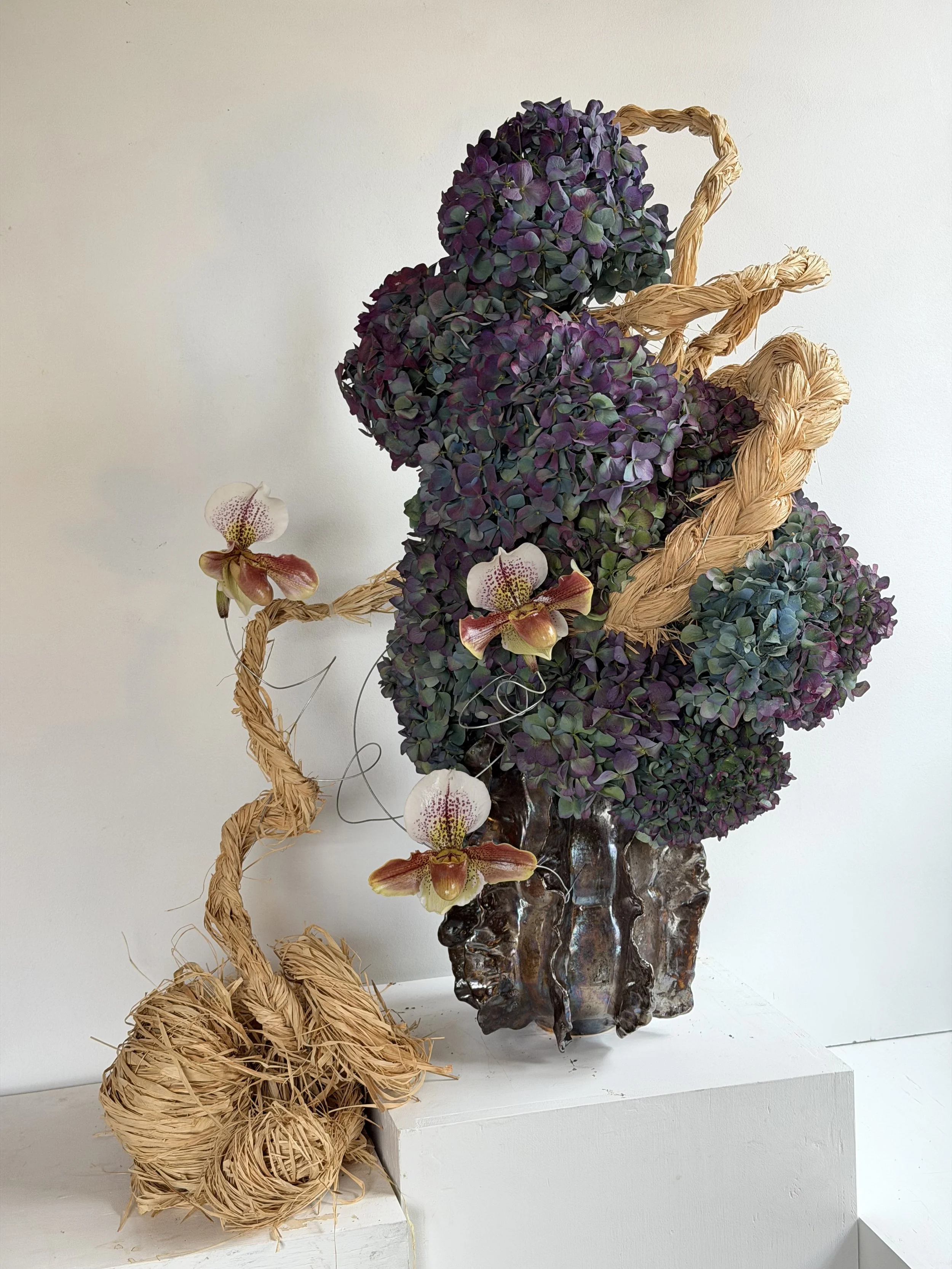 A floral arrangement in a textured ceramic vase featuring purple hydrangeas and pink and yellow orchids, with a decorative twisted straw element arranged around the flowers.