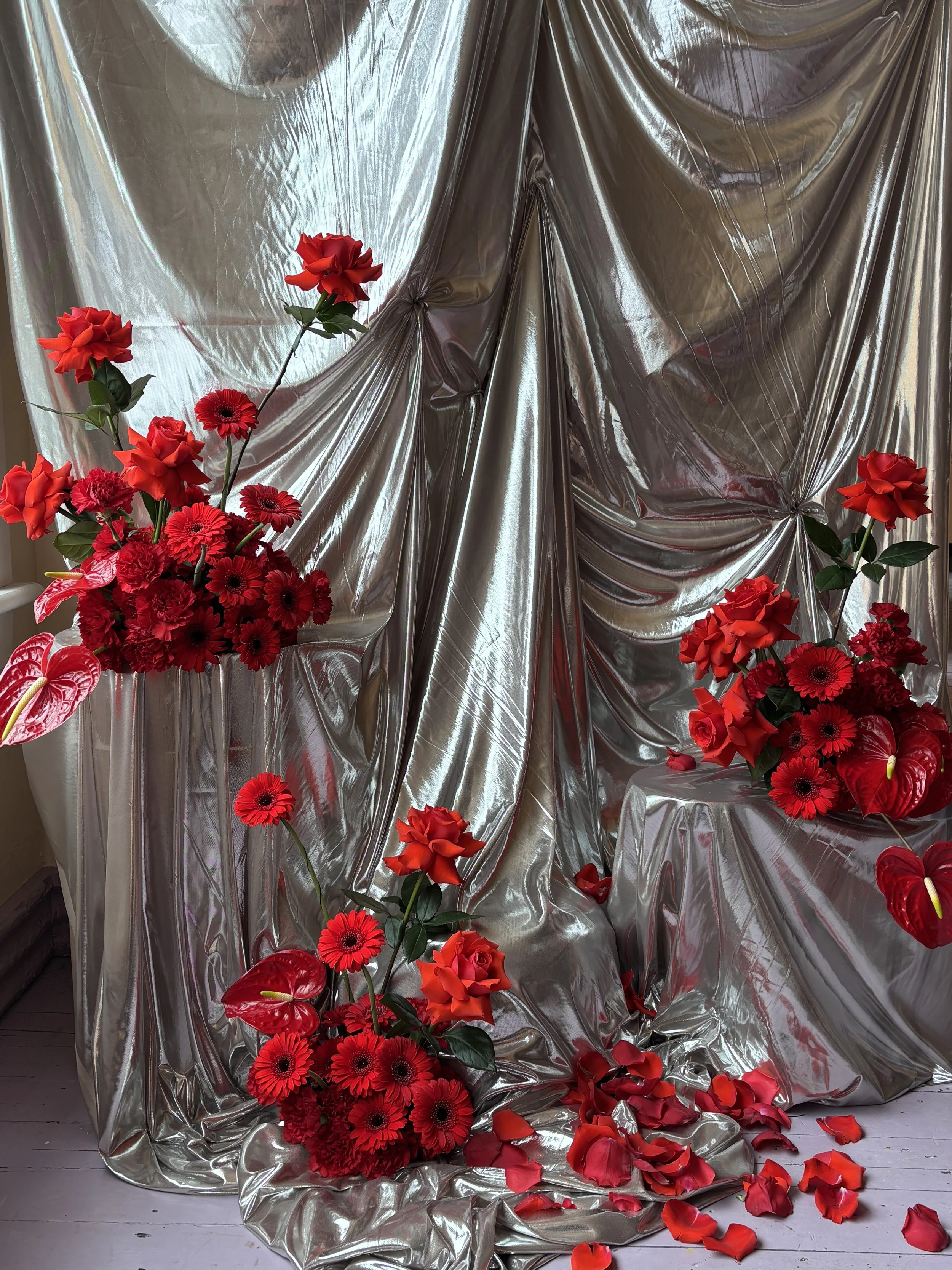 Red flowers, including roses and anthuriums, arranged against a shiny silver draped backdrop with scattered rose petals on the floor.
