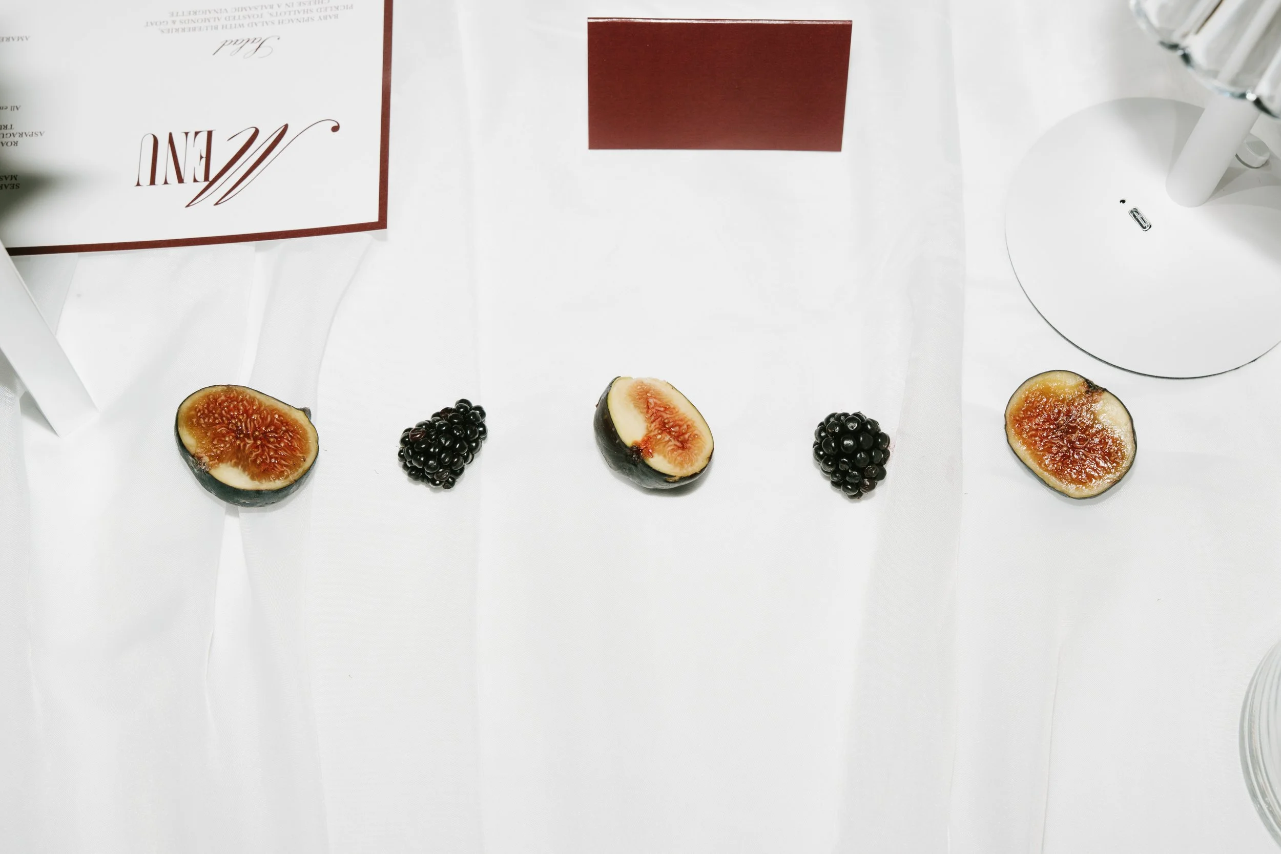 A white table with sliced figs and blackberries arranged in a line, with an open menu and a table lamp visible.