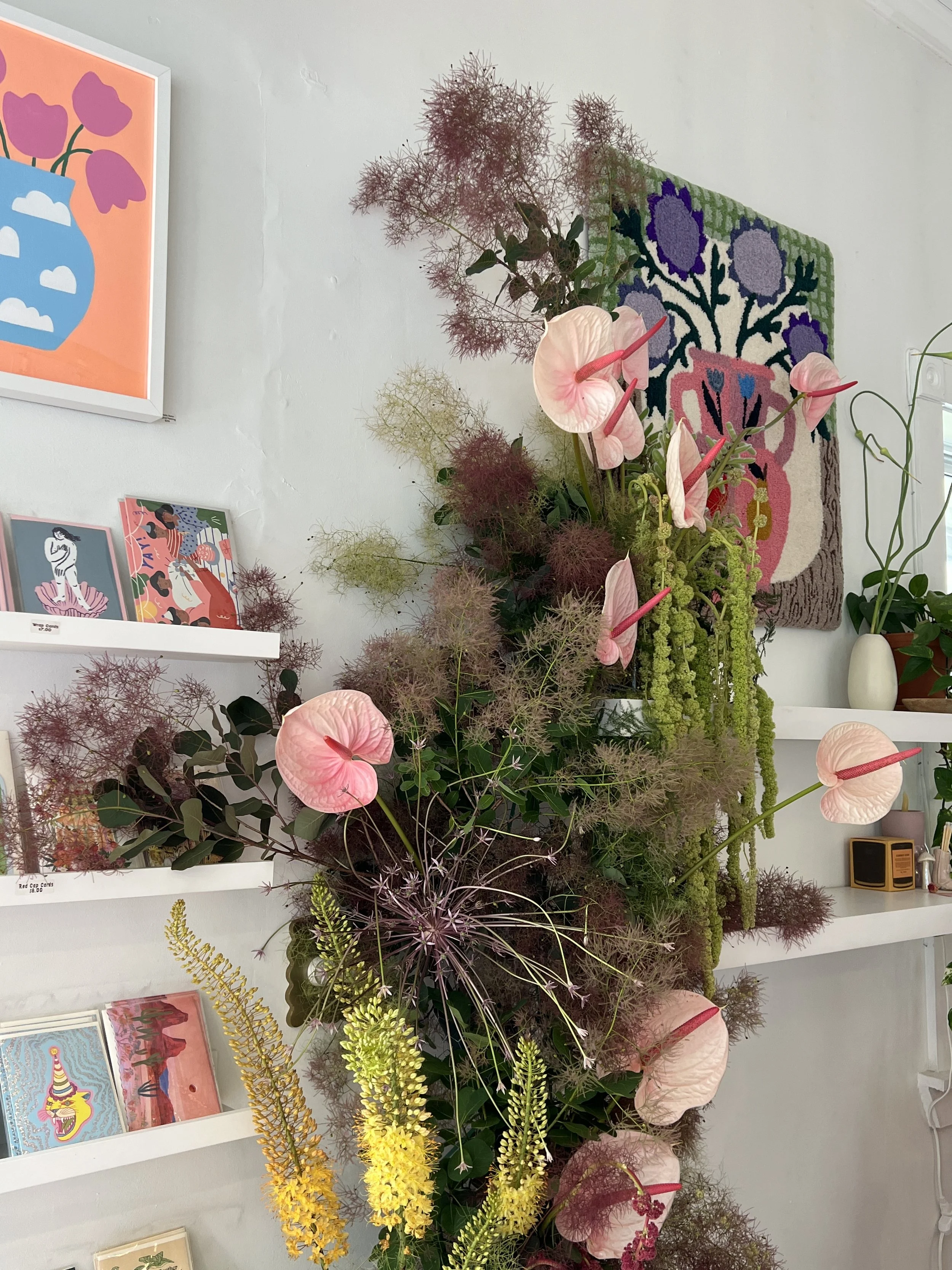 A floral display with pink anthuriums, yellow and purple flowers, and various greenery arranged vertically against a wall, with art and posters on the shelves behind.