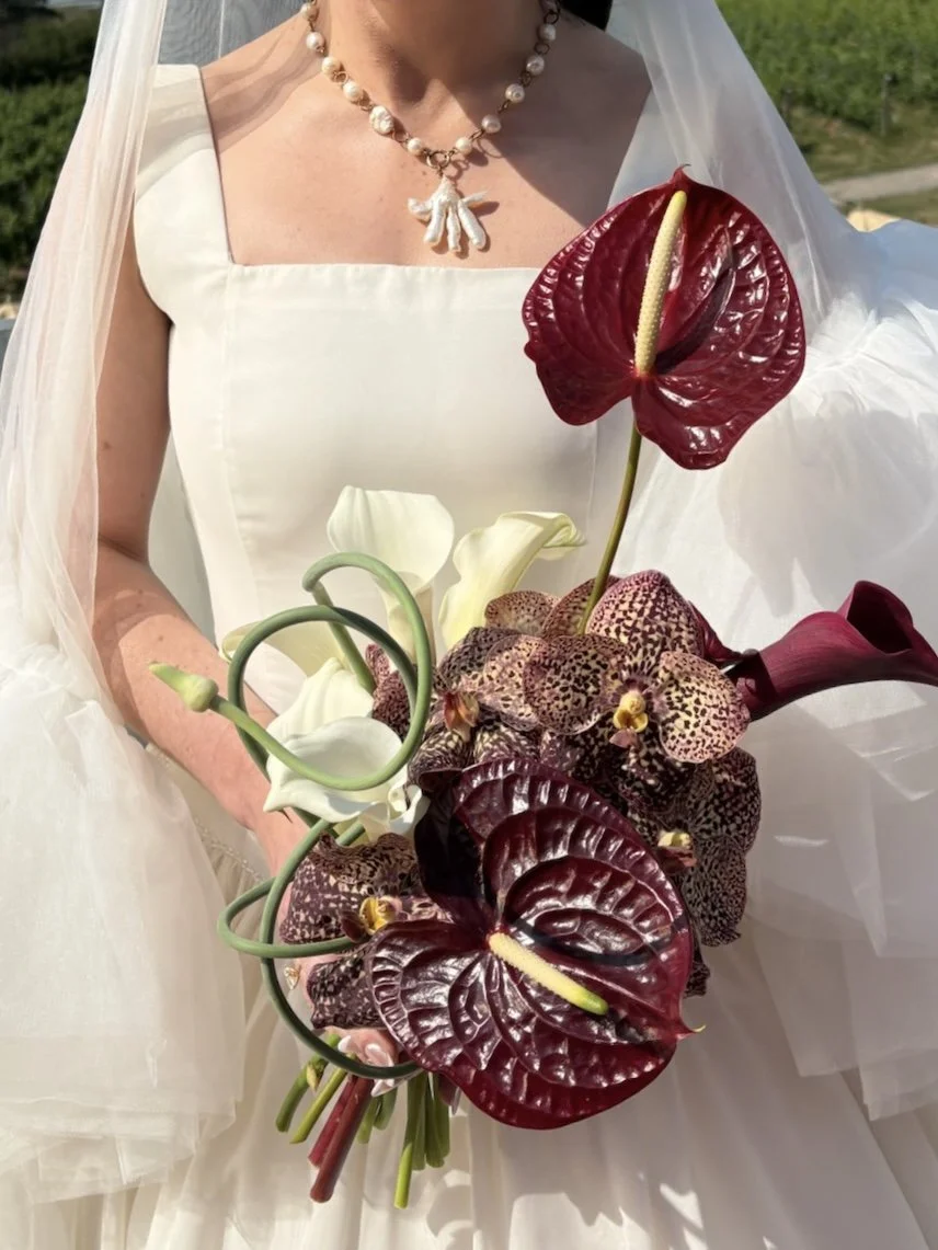 A woman in a white dress holding a bouquet of flowers that includes anthuriums, calla lilies, and orchids. She is wearing a pearl necklace with a pendant.