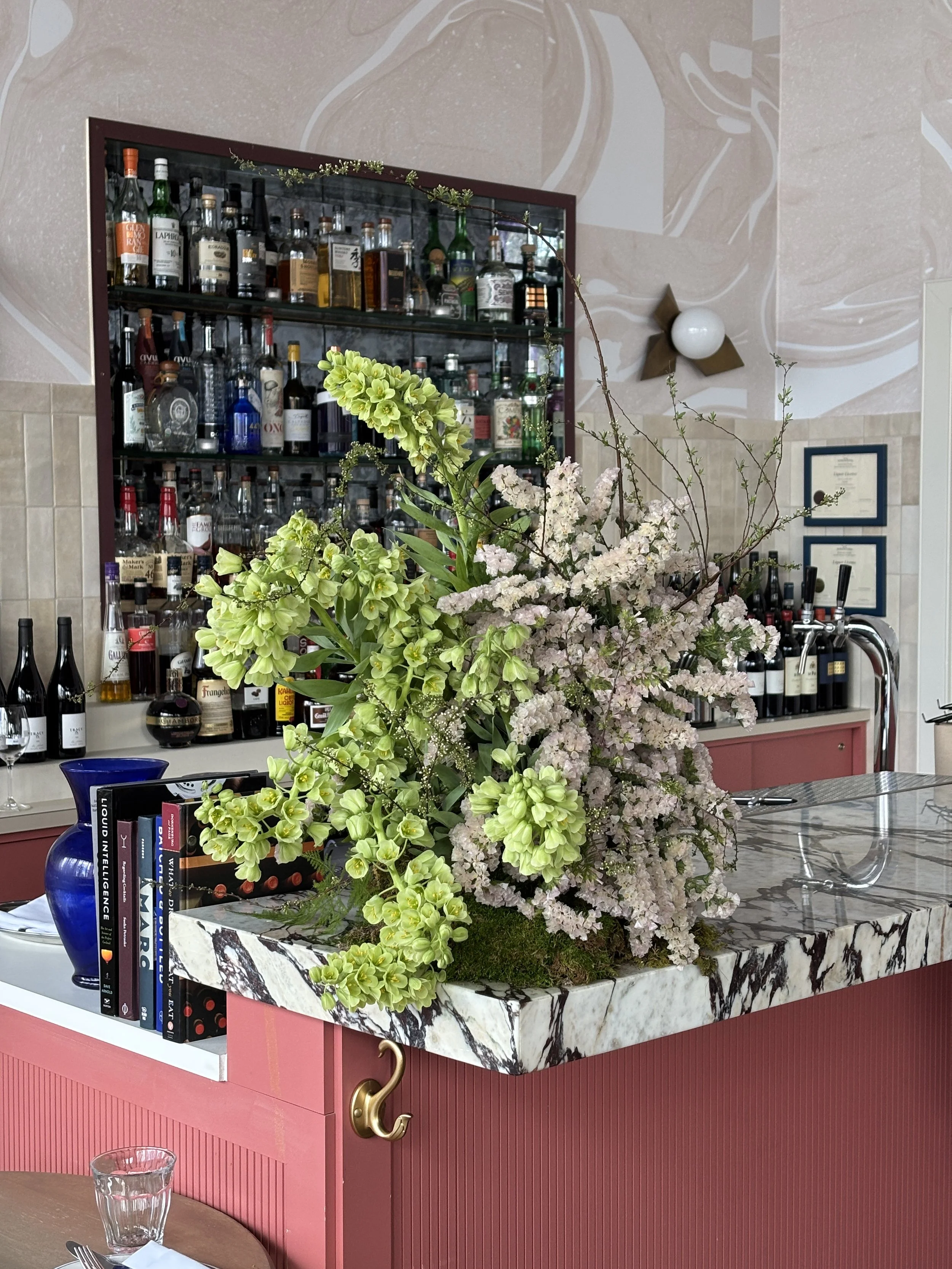 A large floral arrangement with green and pink flowers on a marble countertop in a bar or restaurant setting, with shelves of liquor bottles in the background.