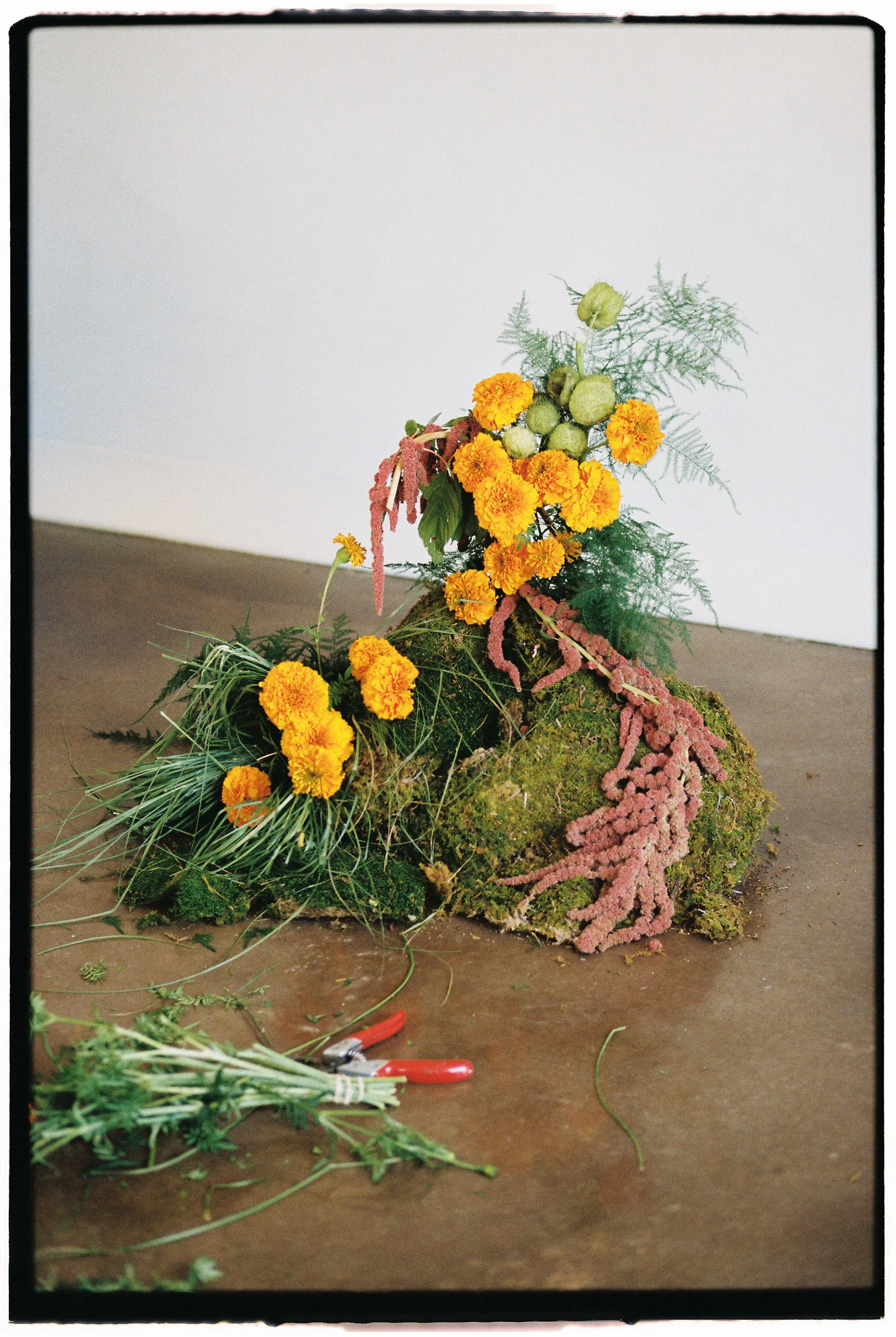 An arrangement of yellow and pink flowers and green plant materials on a moss-covered rock, with gardening tools nearby, set on a brown surface against a plain white wall.