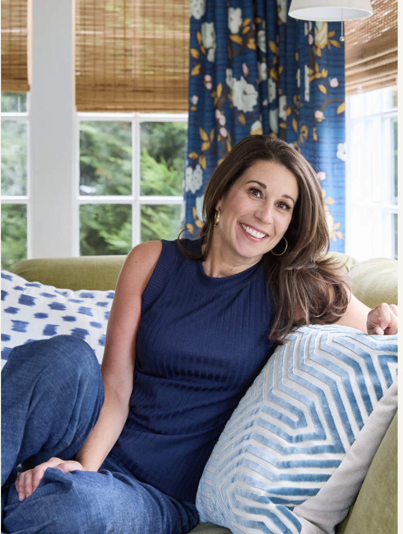 A woman with long brown hair, smiling, sitting on a green couch in a brightly lit room, with large windows and blue floral curtains in the background.