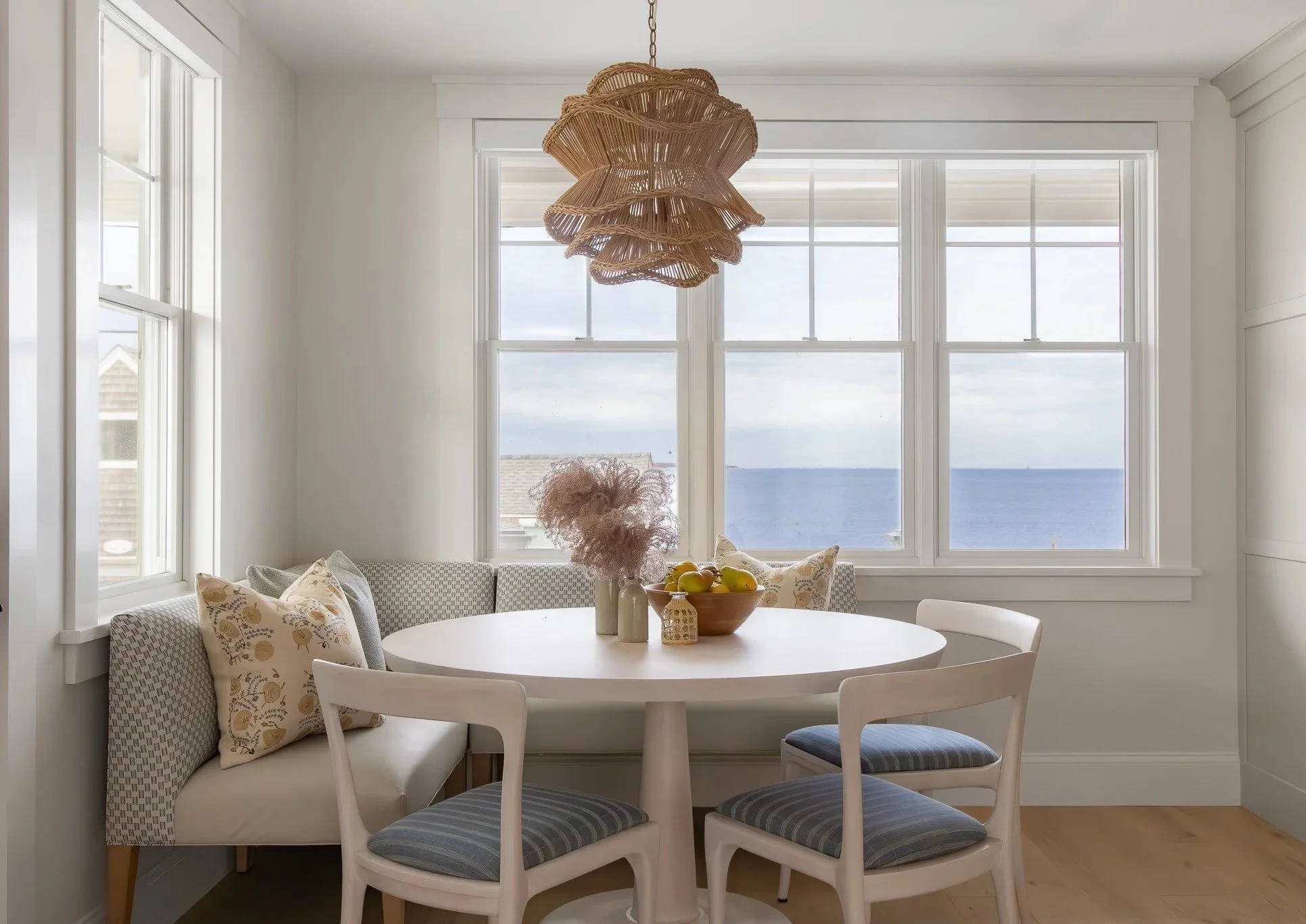 A dining area with a round white table, surrounded by four chairs with striped cushions, and a built-in window seat with floral cushions. A wicker pendant light hangs above the table, and large windows offer a view of the ocean.