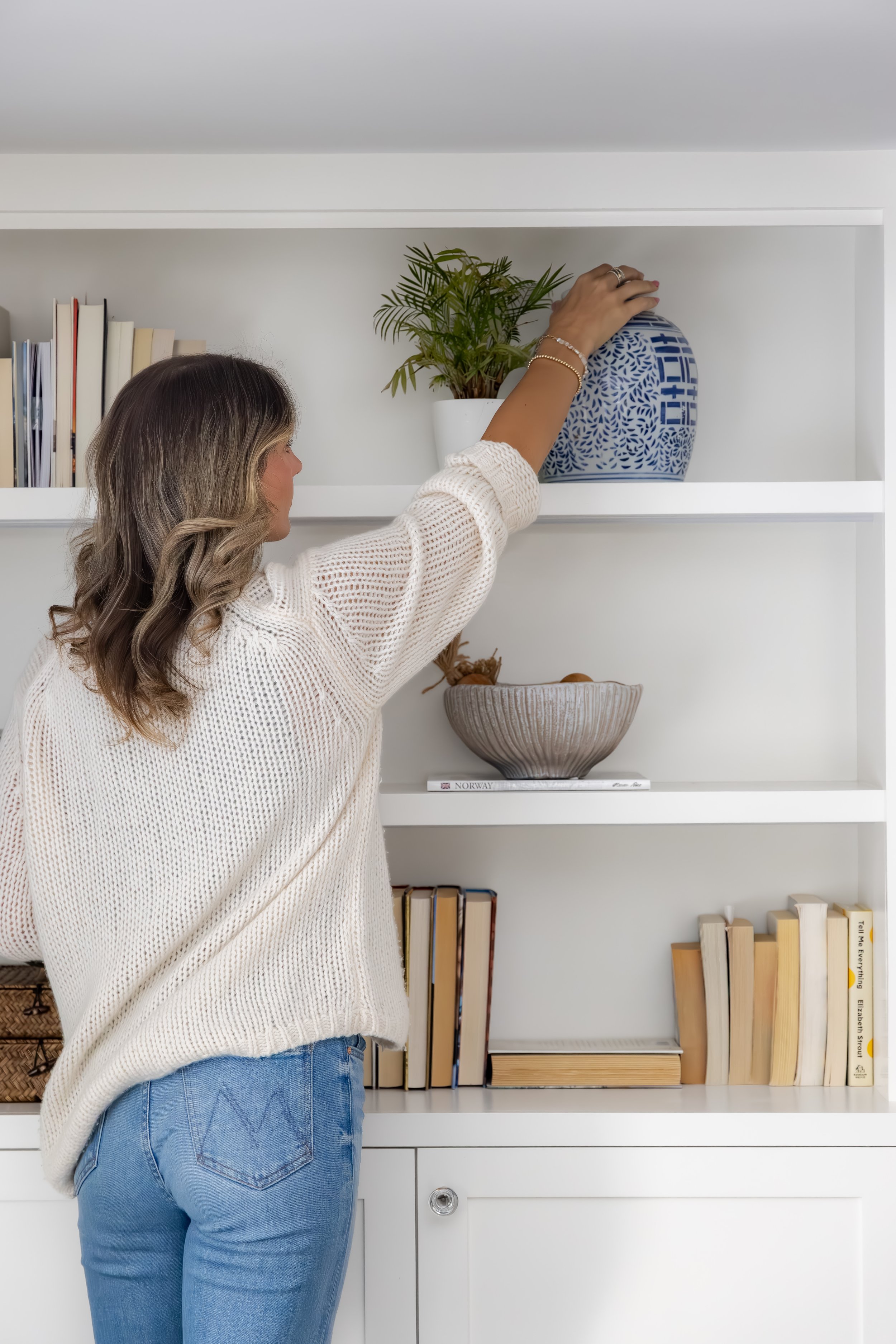 A woman in a loose white sweater and blue jeans arranging a blue and white patterned vase with green plants on a white shelf inside a home.