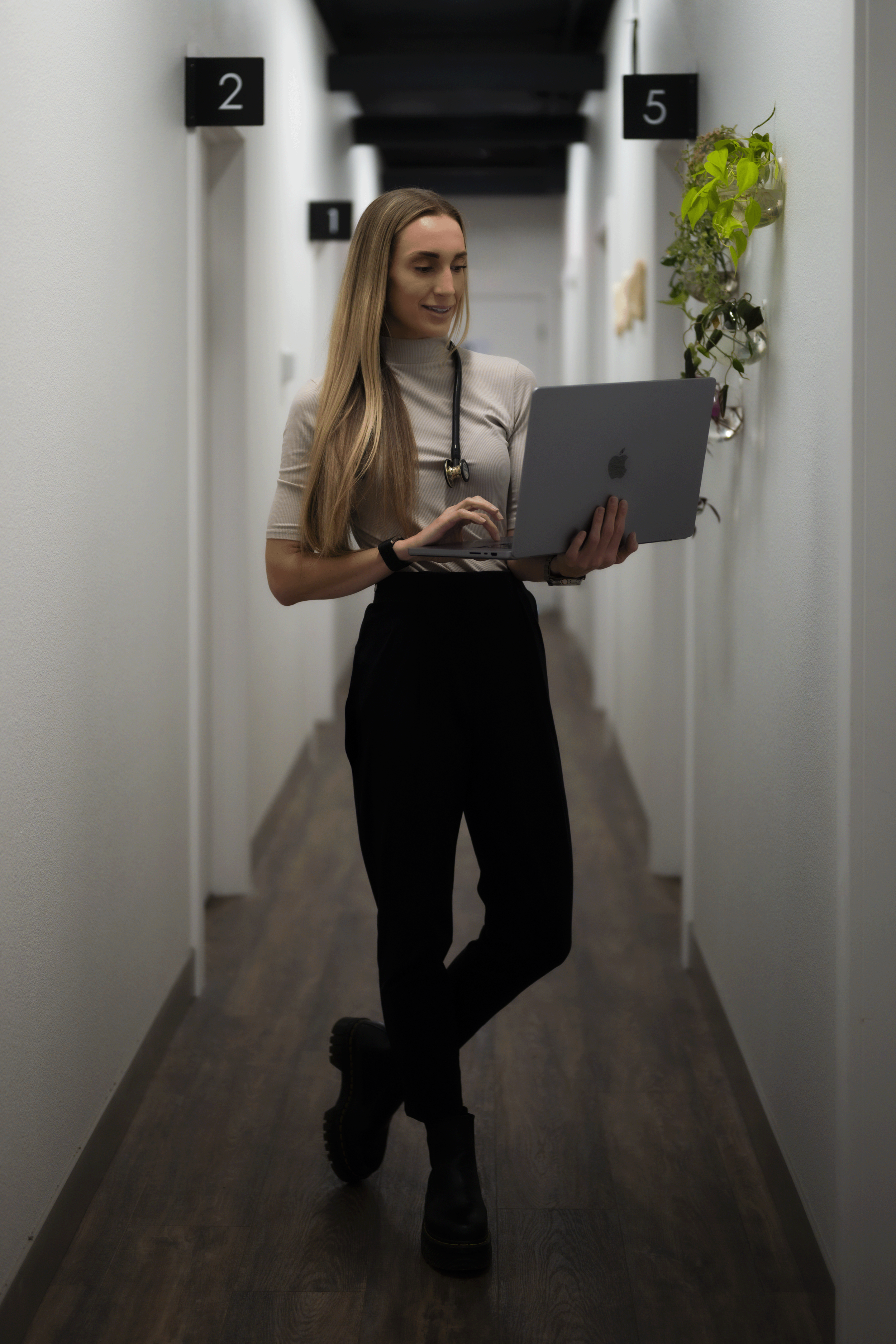 Dr. Erica Strandberg, DO dressed in a beige turtleneck, black pants, and boots, standing in a hallway, looking at a silver Apple laptop.