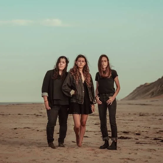 Three women standing on a sandy beach with a mountain in the background