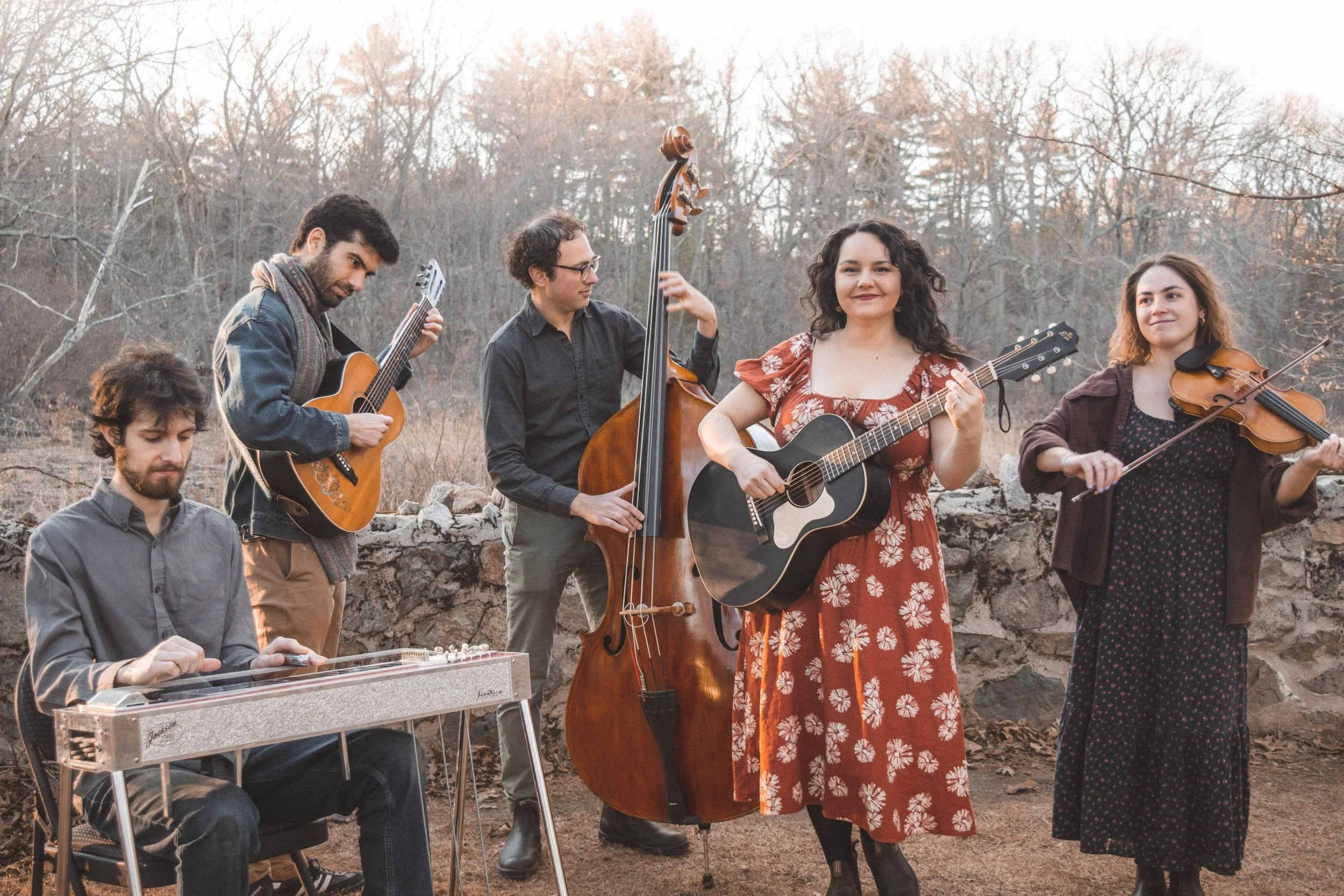 A group of five musicians outdoors playing string instruments and a lap steel guitar. The woman in the center is singing and playing an acoustic guitar.