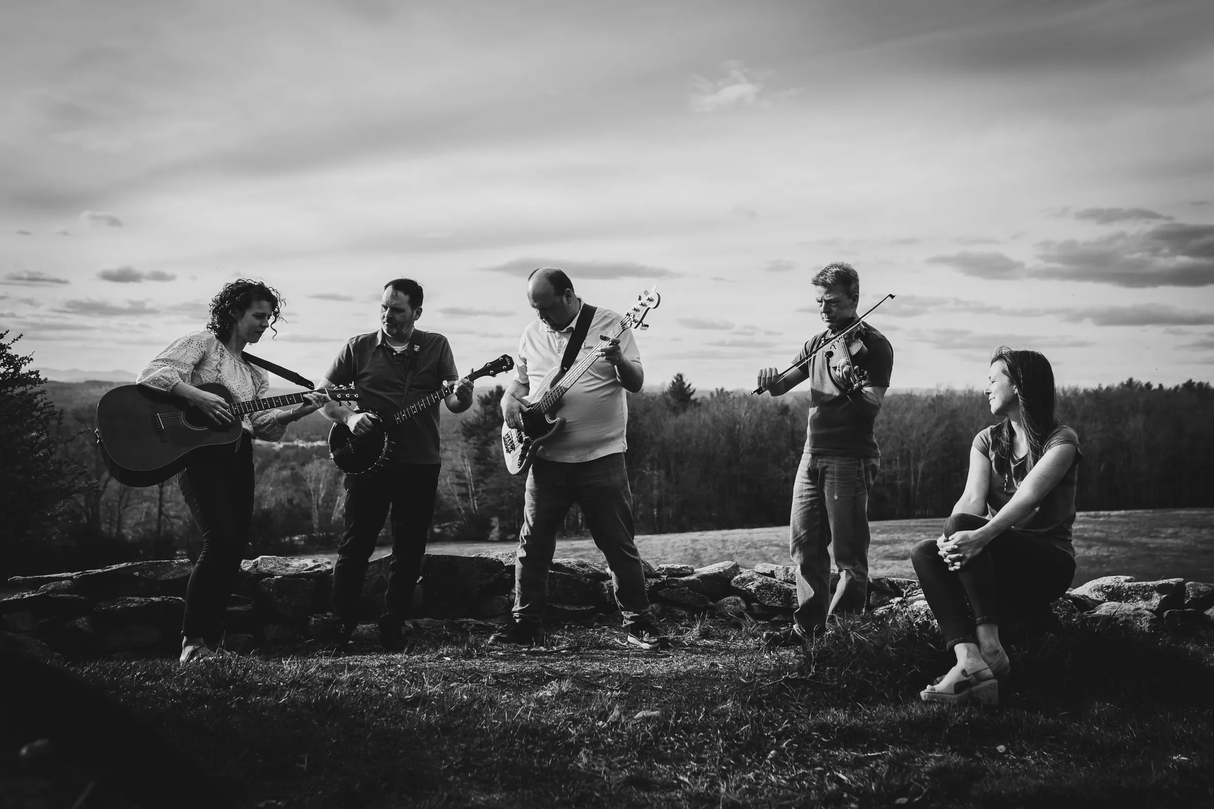 Five musicians playing instruments outdoors during sunsencluding a woman with an acoustic guitar, another with a violin, a man with a bass guitar, and another with a banjo with black and white photo of trees and rocks in the background.