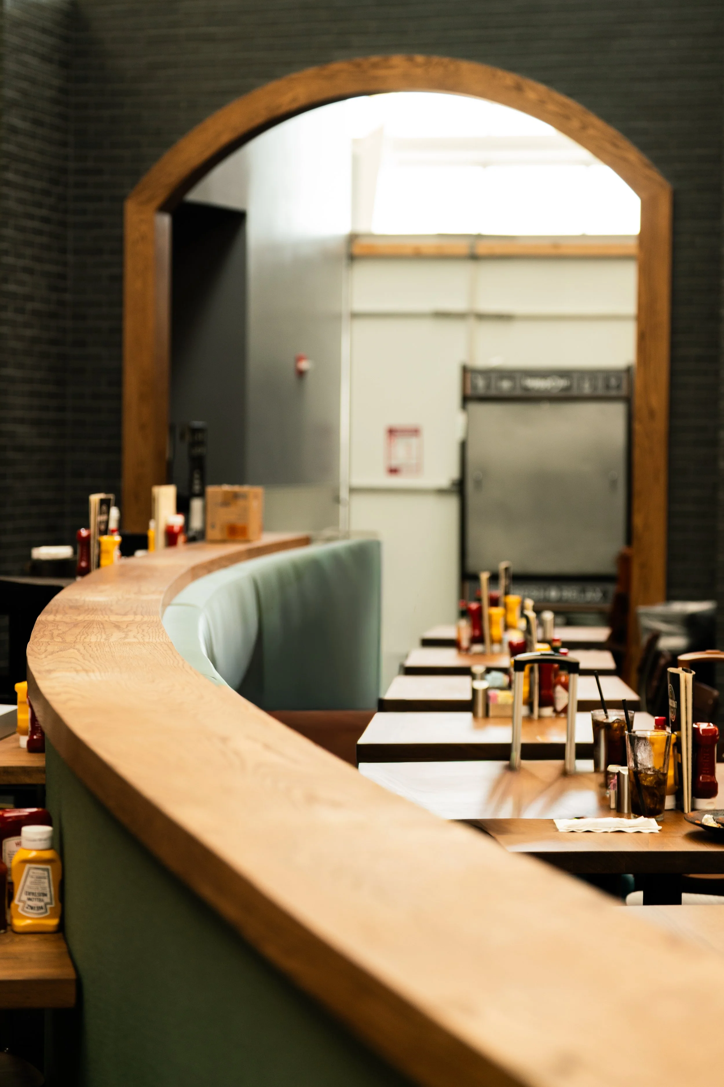 View through a wooden restaurant or cafe window showing tables with condiments and bottles on top, with a soft teal-colored booth in foreground, and a gray door and industrial wall in the background.