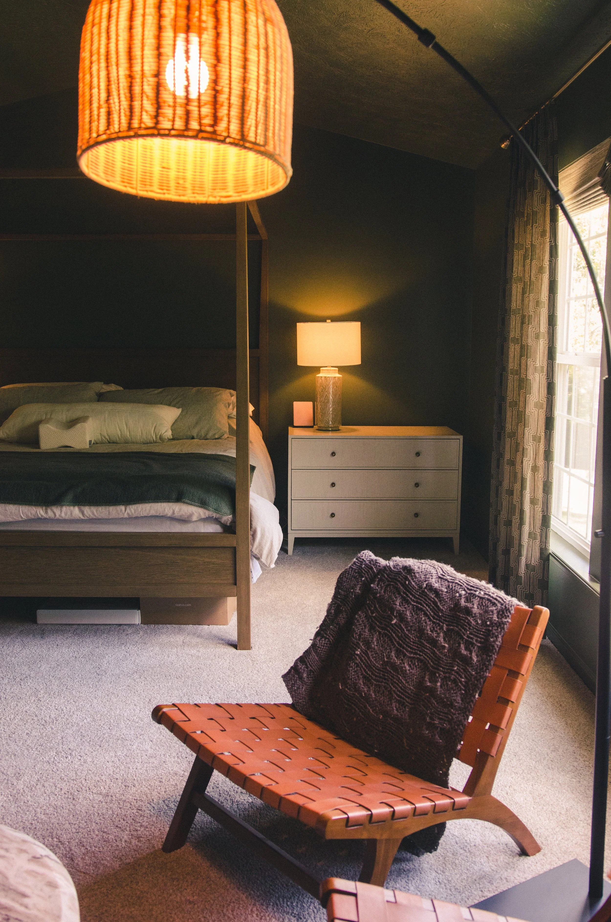 A bedroom with a bed, nightstand, lamp, hanging light fixture, and a chair with a throw blanket and pillow.