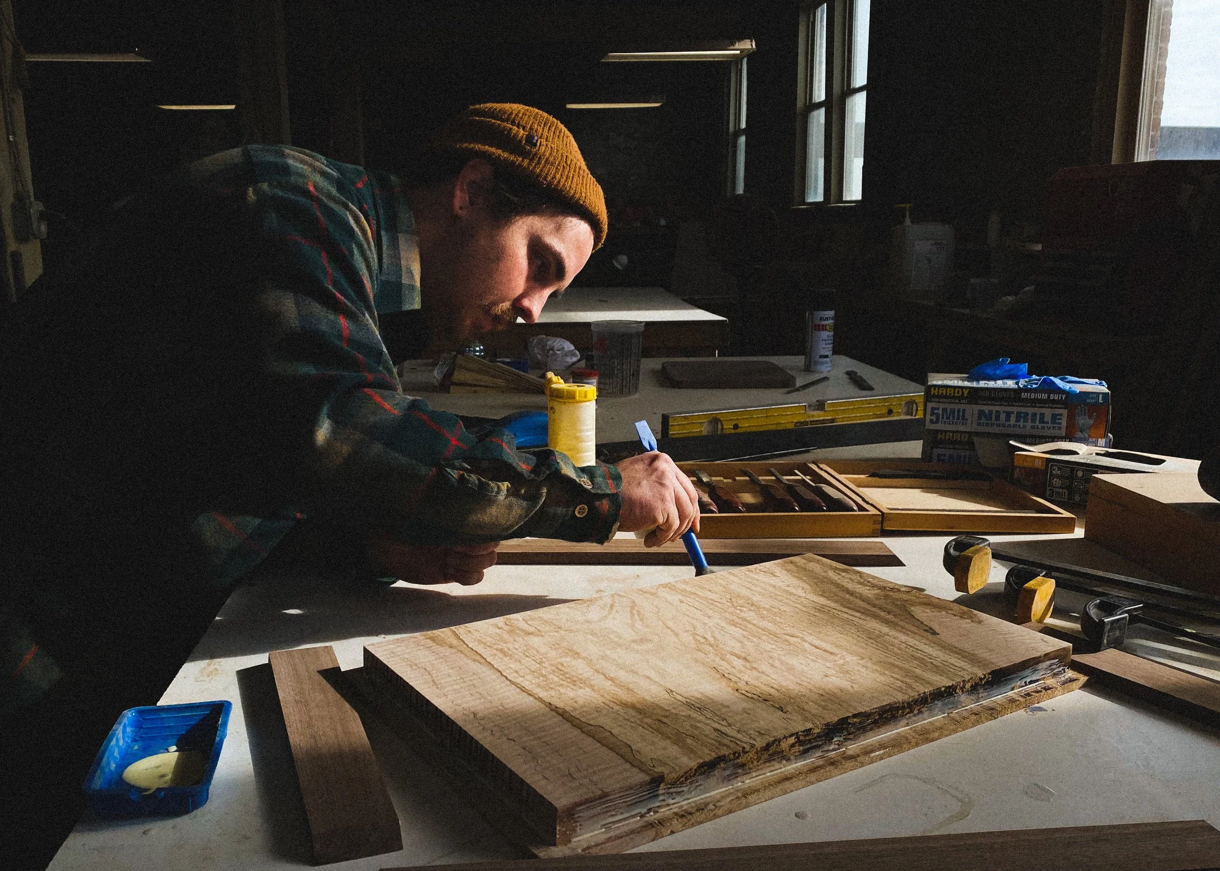 A man wearing a brown beanie and plaid shirt working on a woodworking project in a workshop with natural light coming from a window.