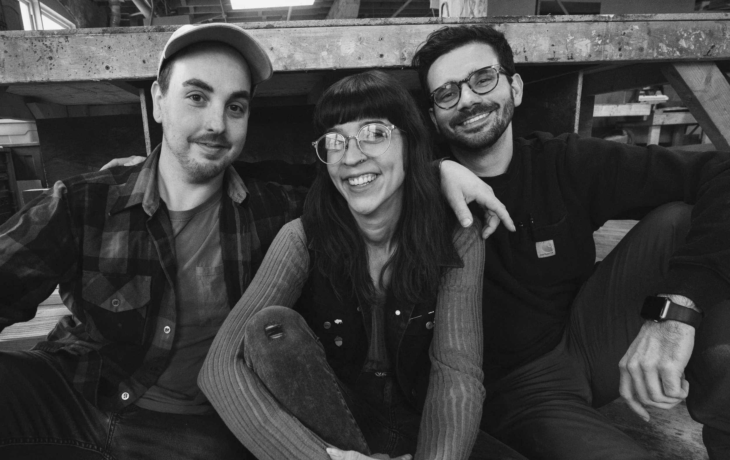 Three smiling friends sitting together, with one woman in the middle and two men on either side, in a casual indoor setting.