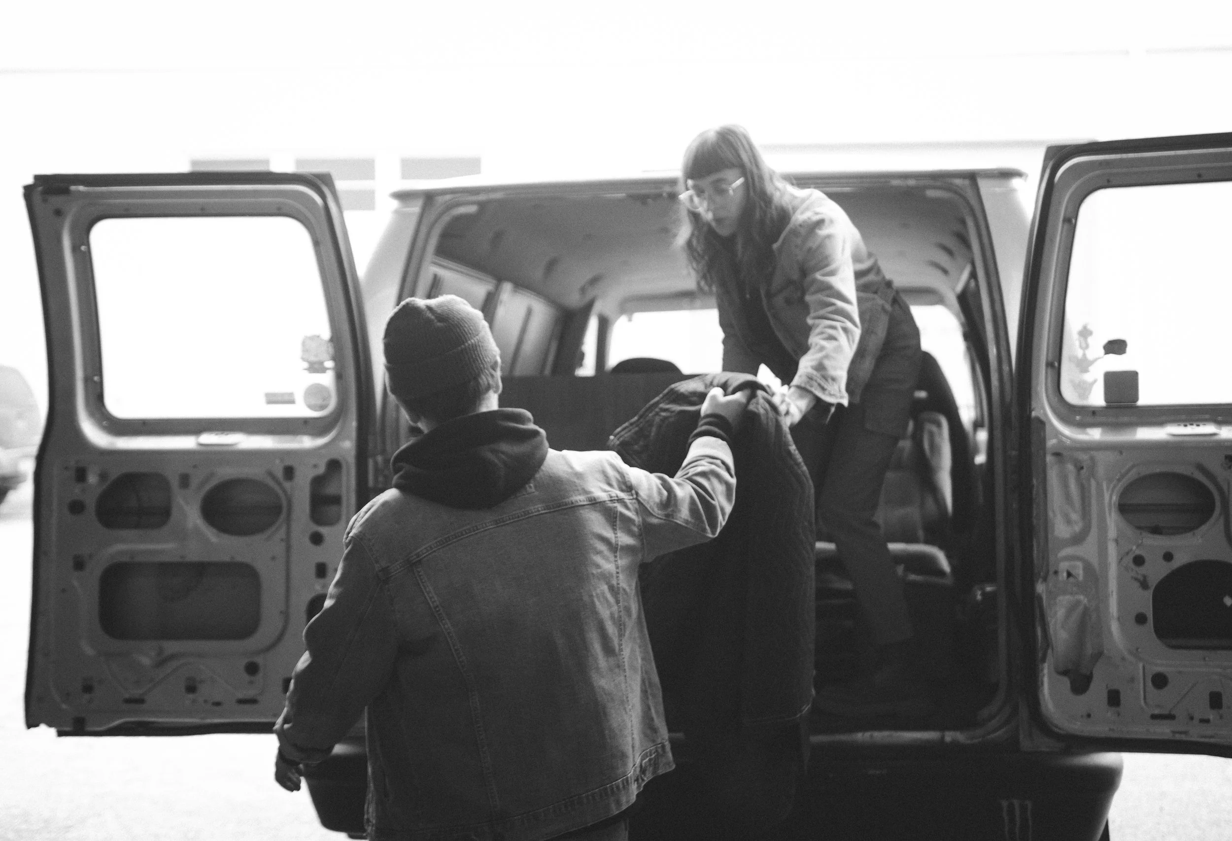 Two people are loading items into the back of a van in a parking lot. One person is inside the van handing items to another person outside.
