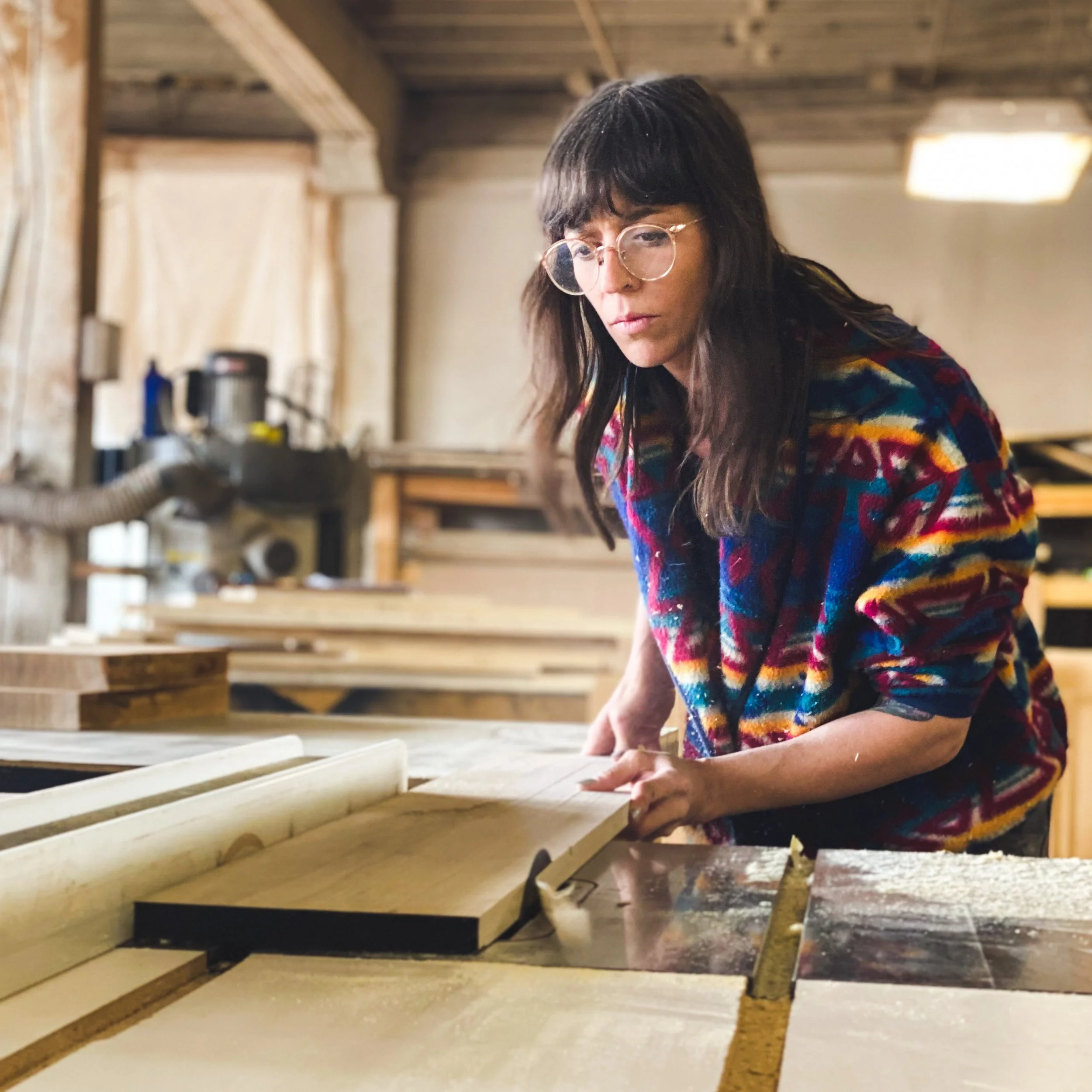 A woman with long dark hair and glasses working with a saw on a piece of wood in a woodworking shop.