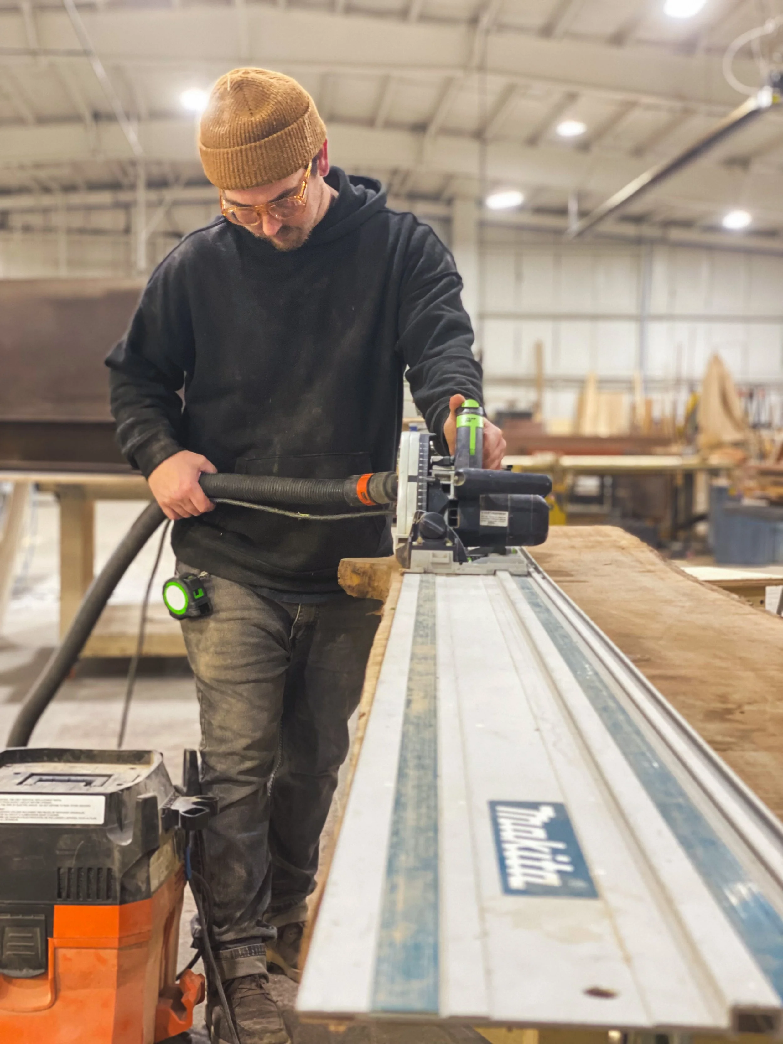 A man in a brown beanie, glasses, and black hoodie using a sanding tool on a wooden board in a woodworking shop.