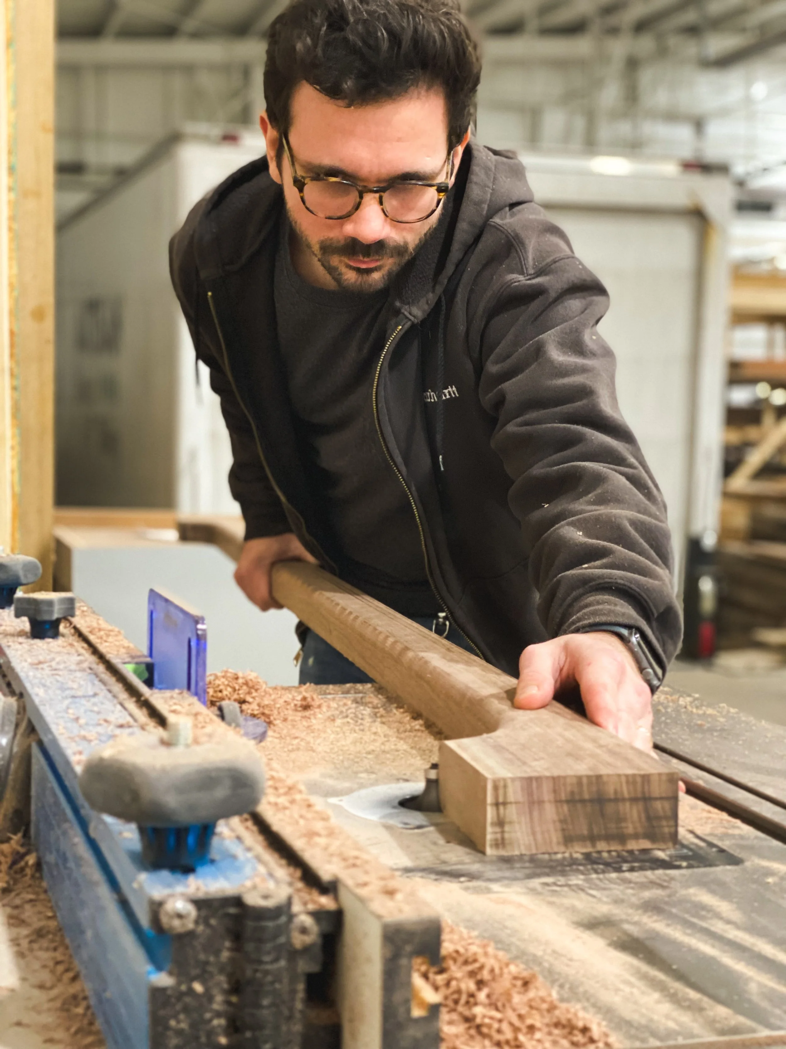 A man in safety glasses and a dark hoodie is working in a woodworking shop, planing a piece of wood on a planer machine.