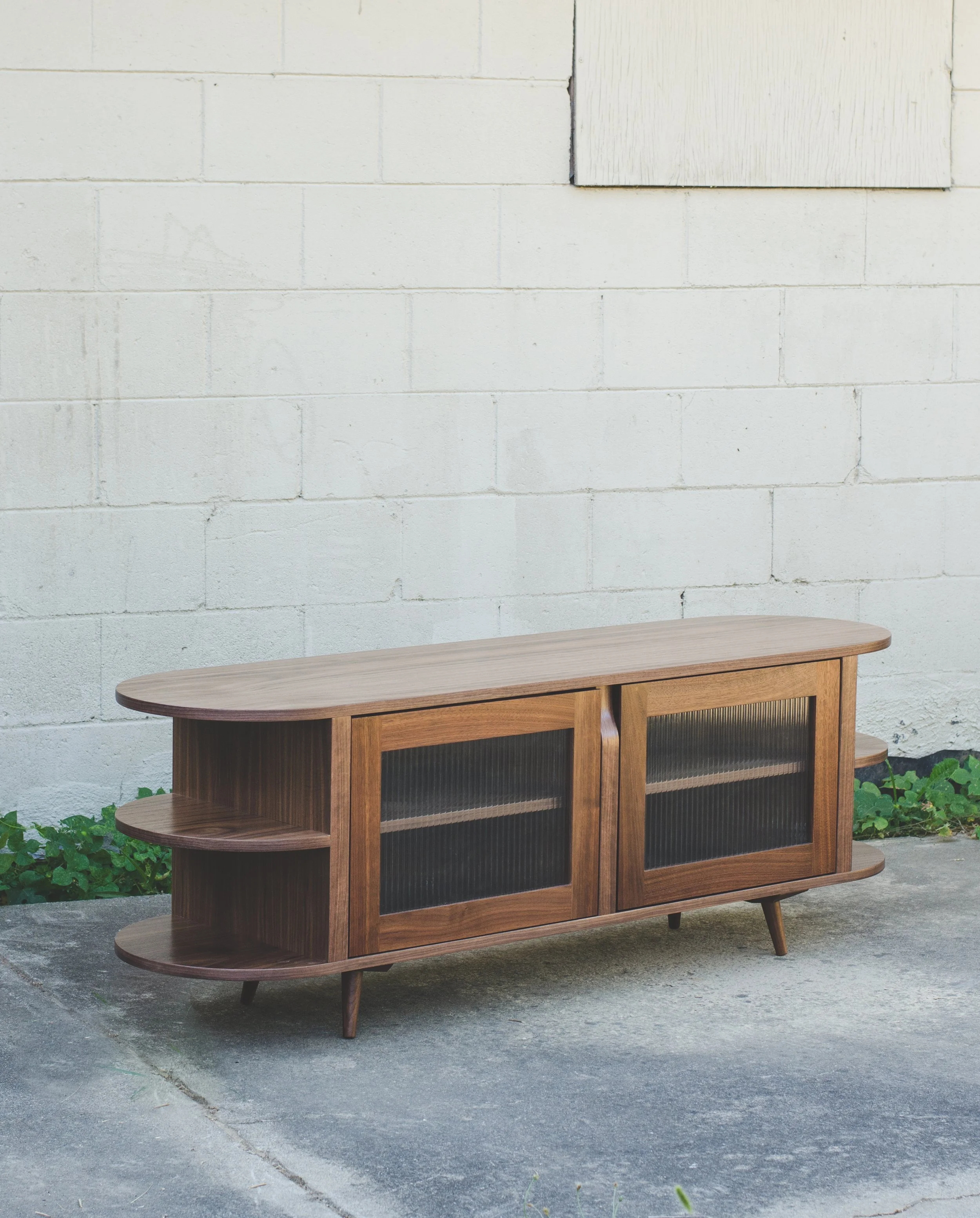 A mid-century modern wooden sideboard with sliding glass doors and rounded edges, set on a concrete floor against a white brick wall with a small window. There are green plants at the base of the wall.